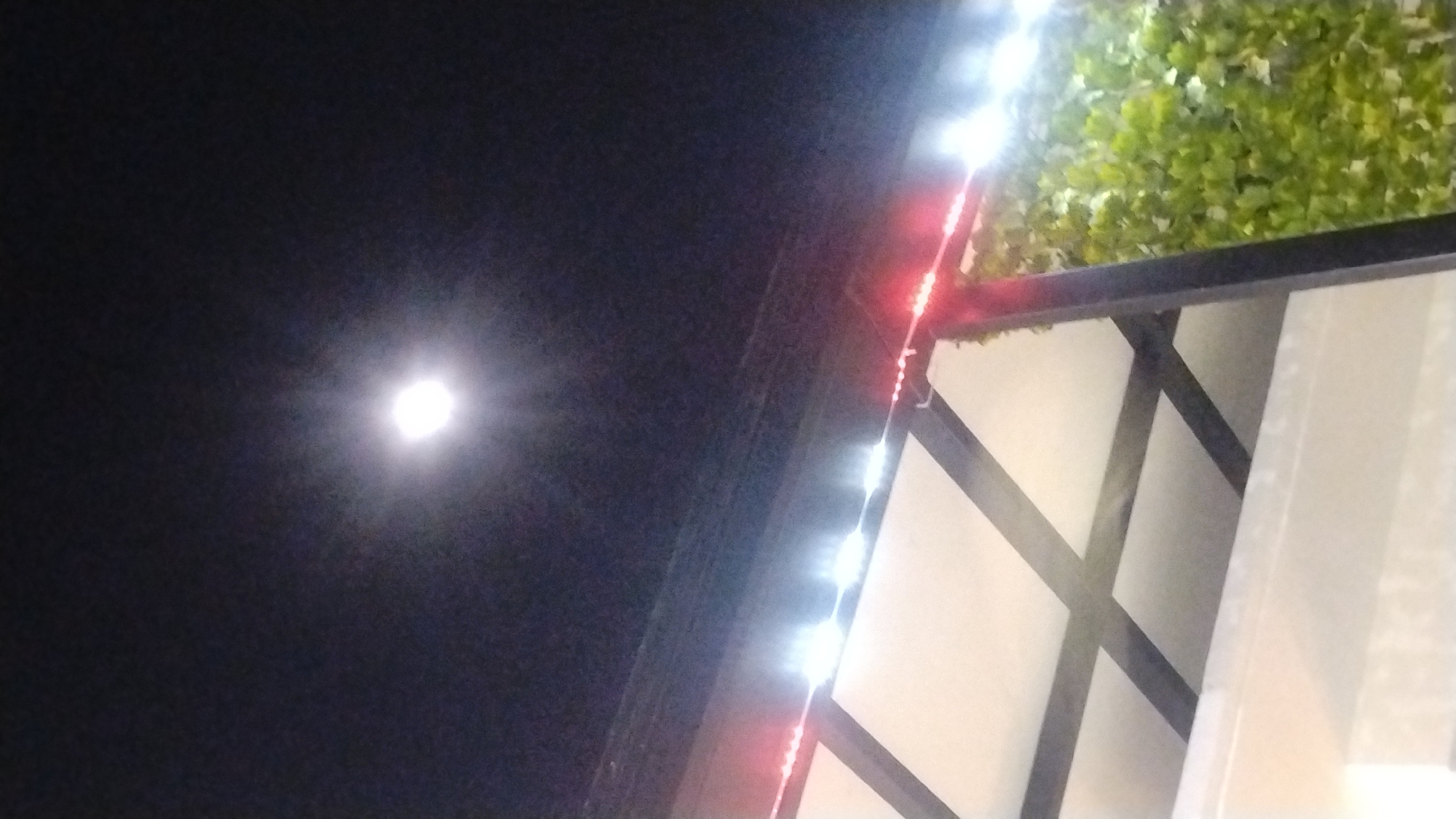 The image shows a night scene with the moon visible in the dark sky. A structure, possibly a roof or awning, is in the foreground, supported by dark beams. String lights, a mix of white and red, are draped along the edge of this structure. To the right, there is a section of lush green foliage, likely plants or vines. The overall impression is that of an outdoor setting at night, possibly a cafe or seating area, illuminated by decorative lights. The image does not contain any people or specific landmarks that would identify its location within the city of Samarinda, Indonesia.