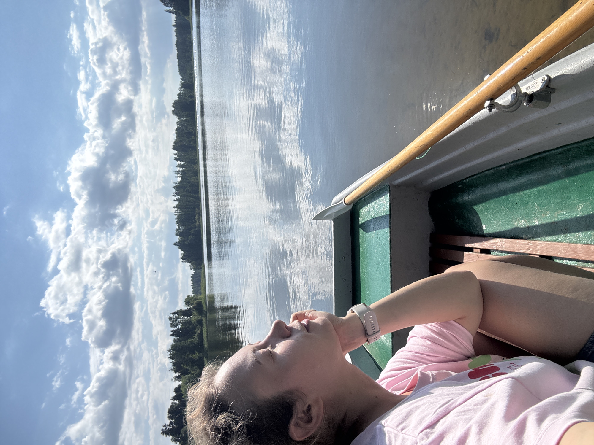 A young woman with blonde hair is sitting in a rowboat on a calm lake, her face turned upwards towards the sky, eyes closed, and right hand supporting her chin. She is wearing a pink t-shirt with a graphic print and a white wristwatch. The rowboat is simple, with green interior seating and wooden benches. A wooden oar rests on the side of the boat. The background shows a reflective lake surface and a distant shoreline lined with trees. The sky is partly cloudy with bright sunlight. This scene is located in Lendži, Latvia, where this type of natural lake environment is common.
