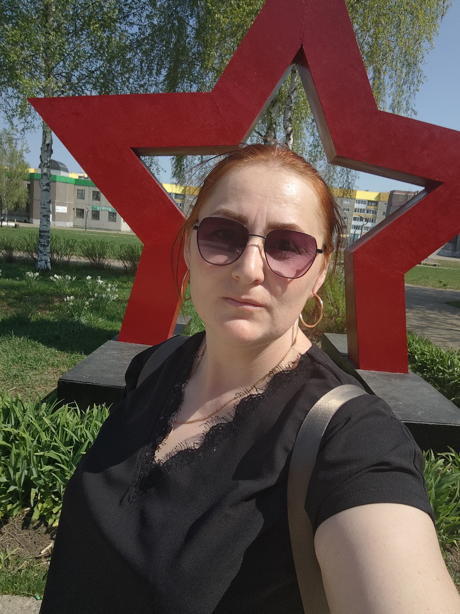 A woman with auburn hair and sunglasses is taking a selfie in front of a large red star in what appears to be a public park or green space in Марковский, Russia. The woman is wearing a black top with lace detailing and gold hoop earrings. She has a strap across her chest, possibly from a bag. Behind her, the red star is mounted on a dark grey base. In the background, there are trees with fresh green leaves, a building with a yellow and green facade, and patches of green grass and white flowers. The sky is clear and blue, suggesting it is a sunny day. The overall atmosphere is casual and outdoorsy.