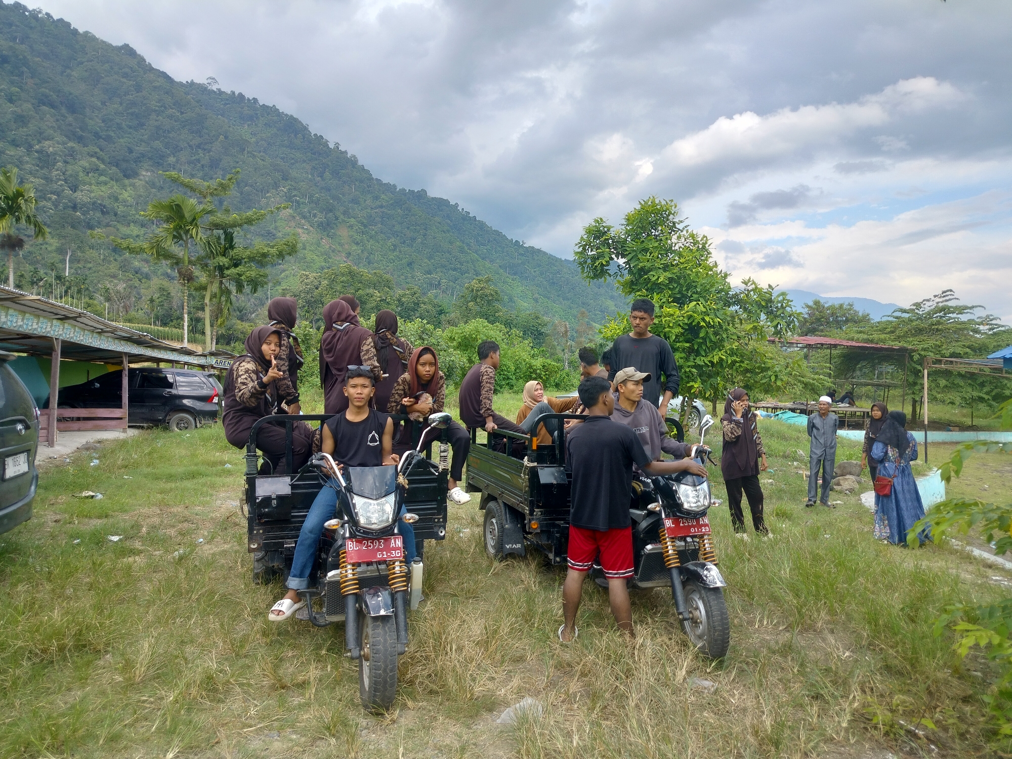 The image depicts a rural scene in Tegarmiko, Indonesia, with a group of people gathered around three small vehicles, possibly for transport or a recreational outing. In the foreground, two motorcycles are parked, one with a young male rider wearing a black sleeveless shirt and blue jeans. He is seated on the motorcycle, which has a license plate reading "BL 2593 AN". To his right, another motorcycle is partially visible, also with a rider.
Behind the motorcycles, a small truck-like vehicle is positioned. Several individuals, mostly young people wearing dark-colored headscarves and clothing, are seated in the back of this truck. A young man in a black t-shirt and red shorts stands beside the truck, facing the viewer. Further in the background, a few adults are visible. One man in a gray suit stands in the middle distance, while a woman in a blue dress and headscarf is on the right.
The environment is characterized by a grassy, uneven terrain with scattered vegetation. In the background, a dense, tree-covered mountainside rises dramatically under a cloudy sky, suggesting a location in a hilly or mountainous region. Structures, possibly shelters or buildings with corrugated roofs, are visible on the left and in the far background. The overall impression is one of a casual gathering in a natural, outdoor setting.