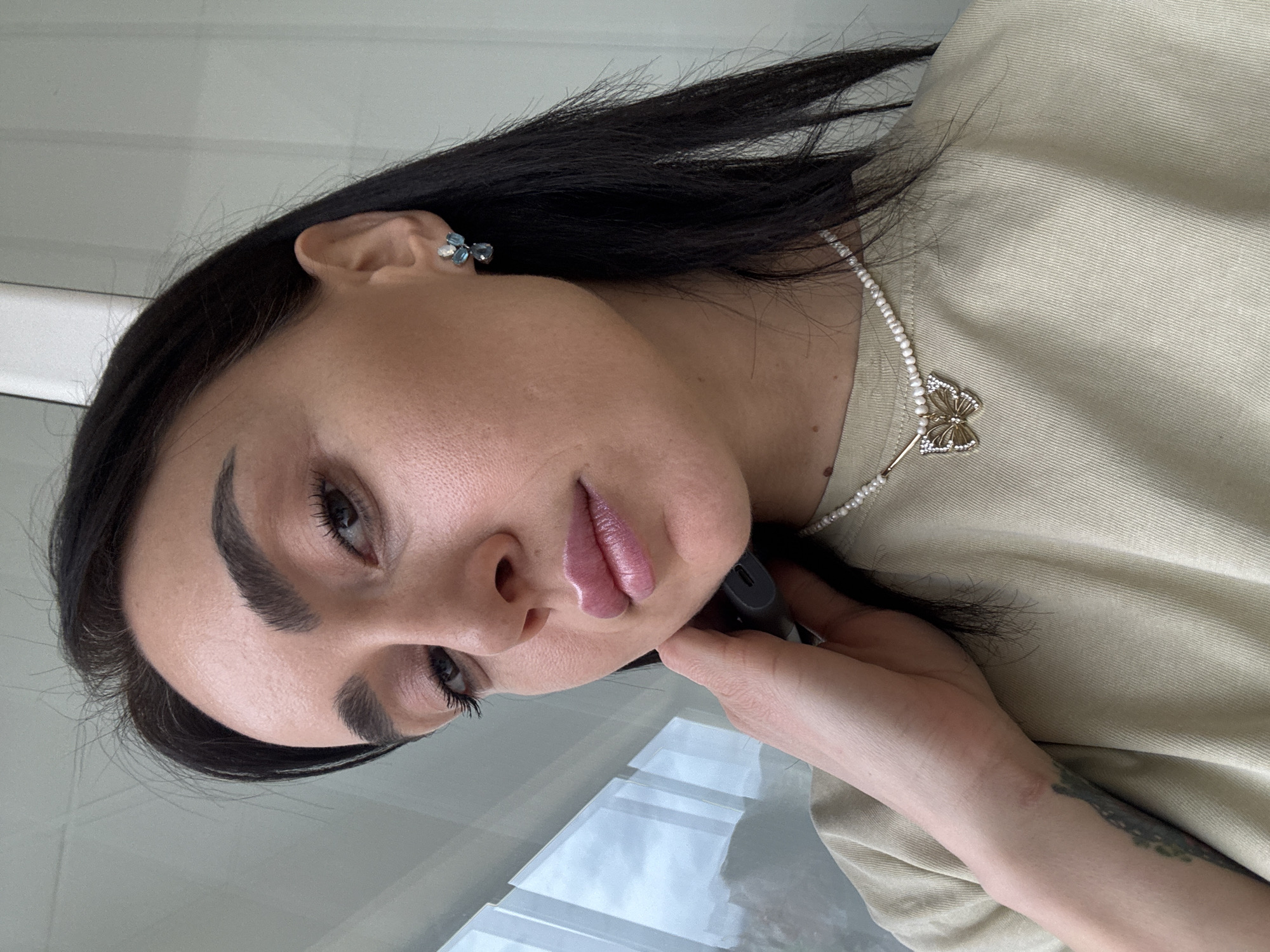 A close-up portrait shows an adult female indoors in Tbilisi, Georgia. She has dark hair, brown eyes, and wears light makeup including lip gloss. Her attire consists of a light beige ribbed t-shirt, a pearl necklace with a gold butterfly pendant encrusted with clear stones, and a cluster earring featuring blue and clear stones in her right ear. Her right hand is positioned near her chin, holding a dark gray, elongated electronic device. A partial tattoo is visible on her right forearm. The background on the left features a window overlooking a cloudy sky and blurred distant structures or terrain, consistent with an urban or semi-urban landscape. The right background is an out-of-focus light-colored wall.