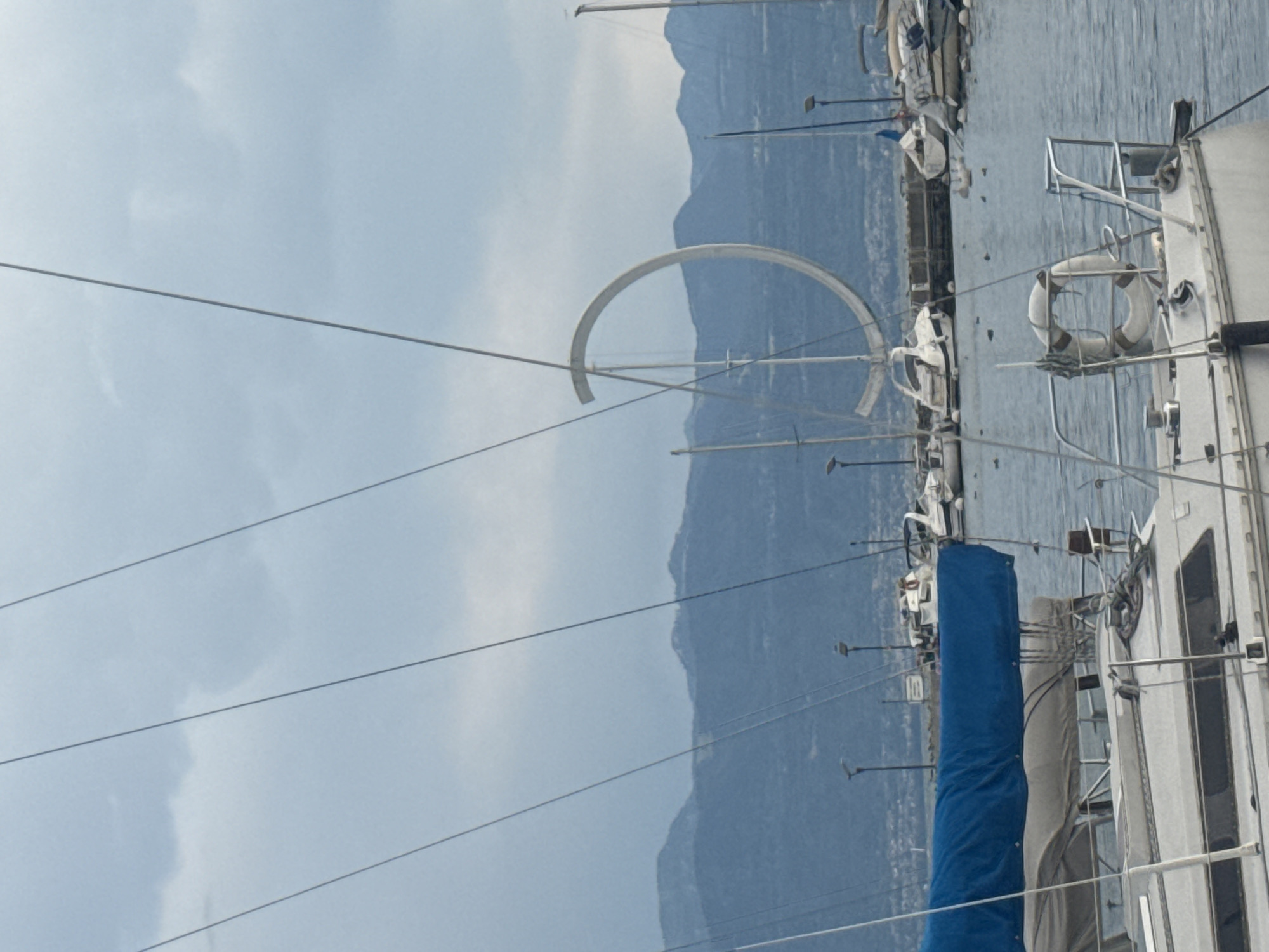 This is a medium-shot, eye-level, vertical photograph taken from a boat, looking across a body of water towards a distant shoreline and mountains. The foreground is dominated by the stern of a white sailboat, with a blue tarp covering its boom and sail. A life preserver ring is mounted on the railing of the sailboat.
In the mid-ground, several other boats are moored, including a white motorboat and various sailboats. A distinctive, large, white ring-shaped sculpture is visible in the sky above the boats, appearing to be suspended or part of a mast structure.
The background consists of a hazy shoreline with buildings and trees, and further in the distance, a range of blue-toned mountains under a cloudy, overcast sky.
The scene is likely the port of Lausanne, Switzerland, given the presence of the "Ouchy Ring" sculpture, a well-known landmark in the Ouchy district of Lausanne. The water is calm, and the overall atmosphere is muted due to the cloudy weather. There are no people clearly visible in the image.