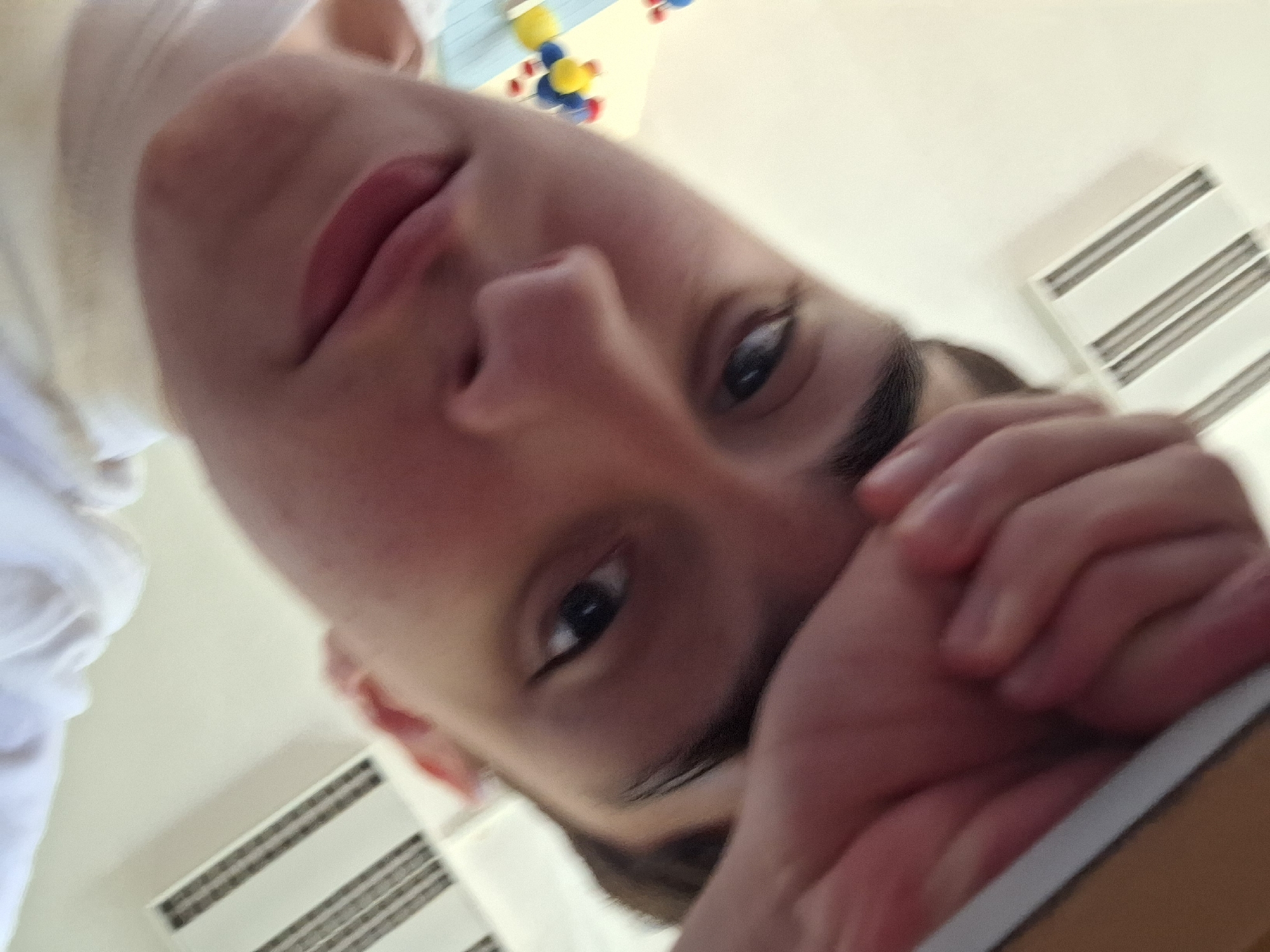 The image is a selfie taken from a low angle. A young person's face, with dark hair and eyebrows, is visible in the foreground. Their left hand is raised to their forehead. In the background, to the left, are some colorful, spherical objects hanging from strings, resembling a mobile or toy. Further to the left and right, there are white rectangular objects with horizontal black stripes, possibly air vents or decorative elements on a wall. The overall lighting is bright and appears to be indoors. The subject's face is partially obscured by their hand and the angle of the camera. There is no indication of the city of Varna, Bulgaria, or any specific location within it in the image.