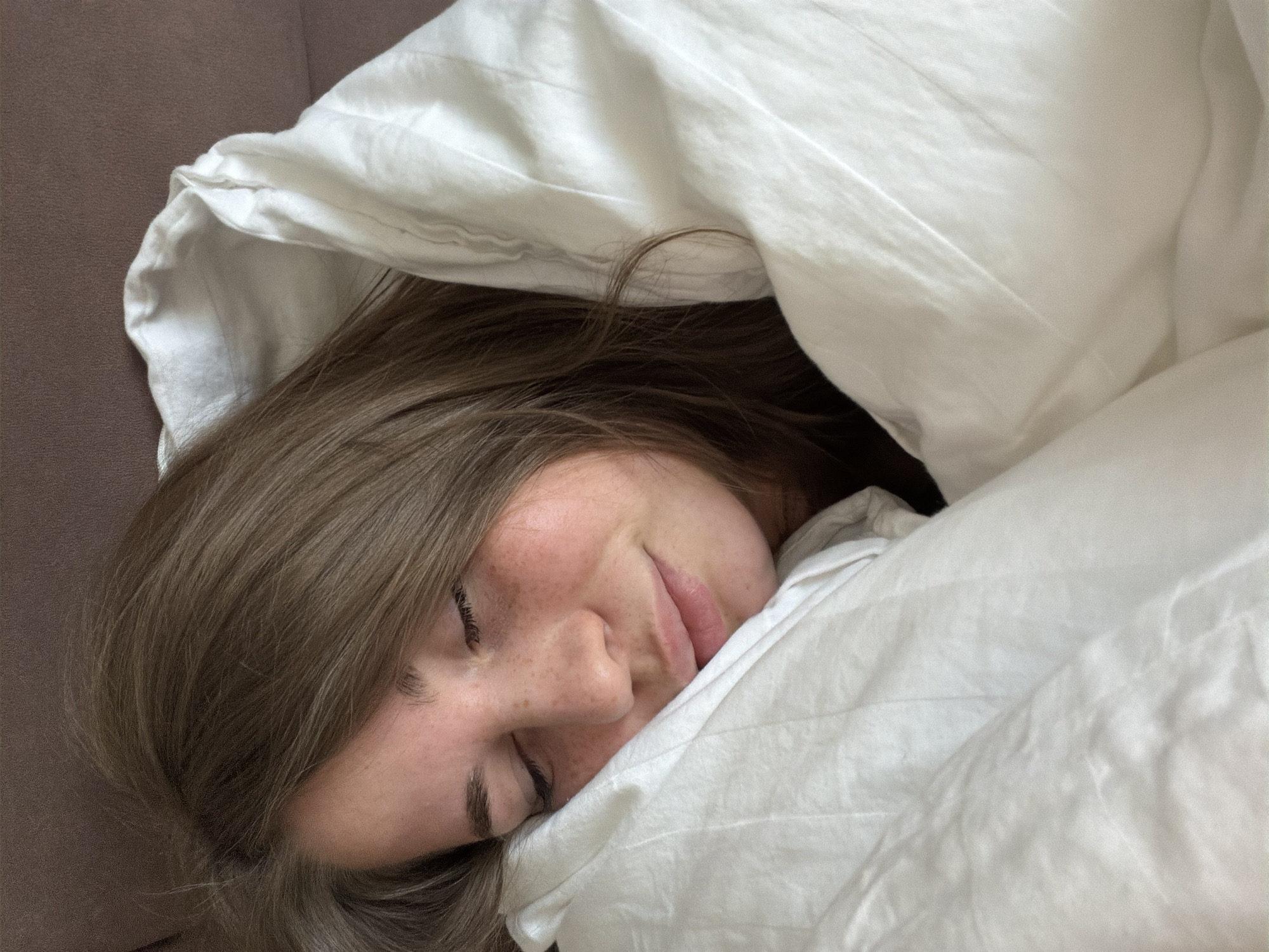 A young adult individual, gender presenting as female, is depicted in a close-up, high-angle portrait, appearing to be asleep or resting. Her eyes are closed, and her facial expression is serene. Light brown hair frames her face, partially obscuring her forehead and extending onto the white bedding. Visible freckles are present across her nose and cheeks, with a slight reddish flush on her skin. Her lips are lightly parted. She is enveloped in a white duvet or blanket, which is pulled up around her neck and shoulders, and a white pillow or pillowcase supports her head. The white fabric displays soft creases and folds. The background consists of a blurred, solid reddish-brown surface, consistent with furniture or a wall, in the upper portions of the image. The scene is set in an indoor environment, location Unknown, Unknown.