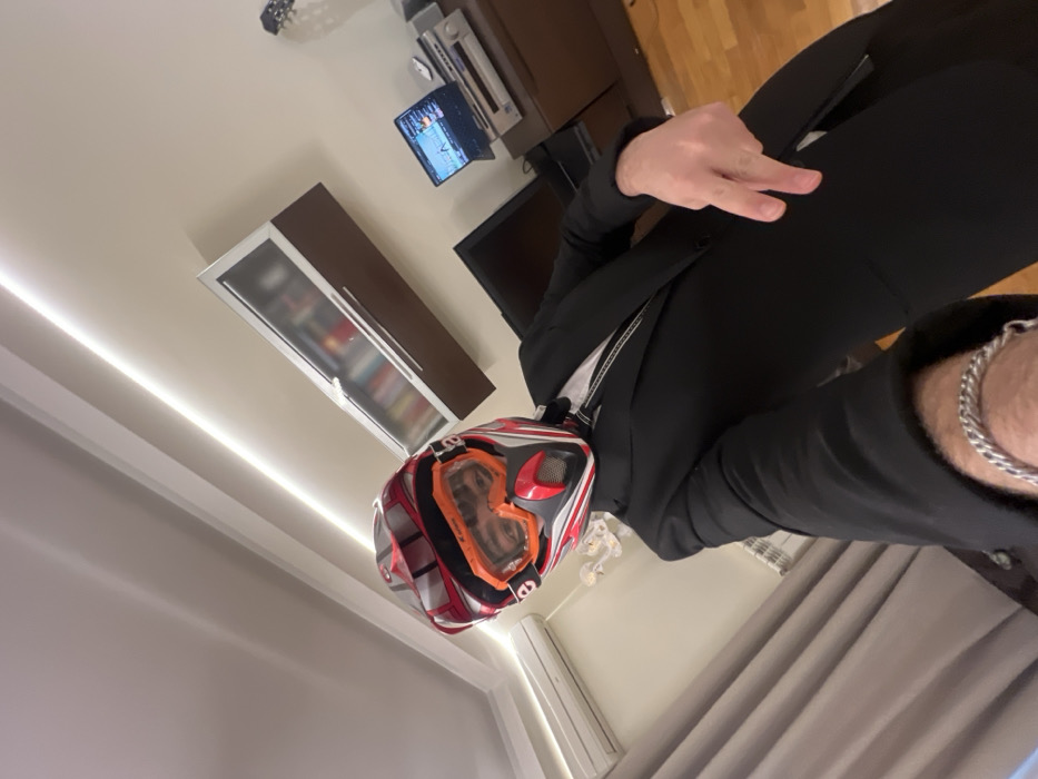 A person in a black suit is wearing a motocross helmet with orange goggles. They are making a peace sign with their right hand. The background shows a room with a brown cabinet, a bookshelf with books, a television, and a wooden floor. A silver chain bracelet is visible on the person's left wrist. The image appears to be taken from a low angle, tilted slightly.