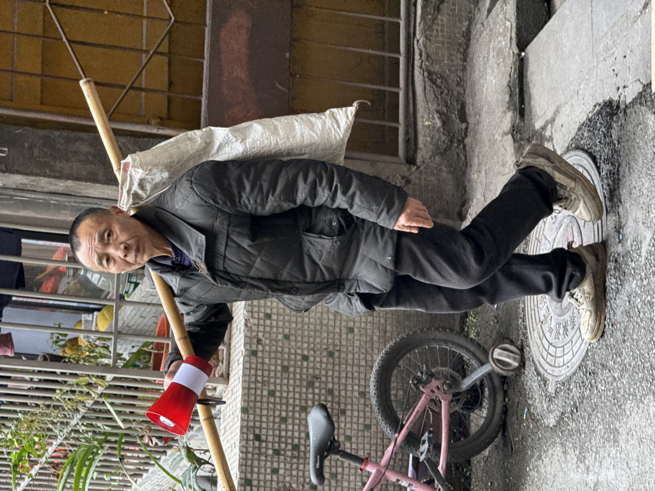 An elderly Asian man stands beside a pink bicycle. He wears a black quilted jacket, dark pants, and worn sneakers. He balances a bamboo pole on his shoulder, carrying a white bag. A red and white megaphone hangs from the pole. He stands on a concrete slab with circular markings, next to a tiled wall. A building with a barred window and potted plants can be seen in the background. The image is slightly tilted.