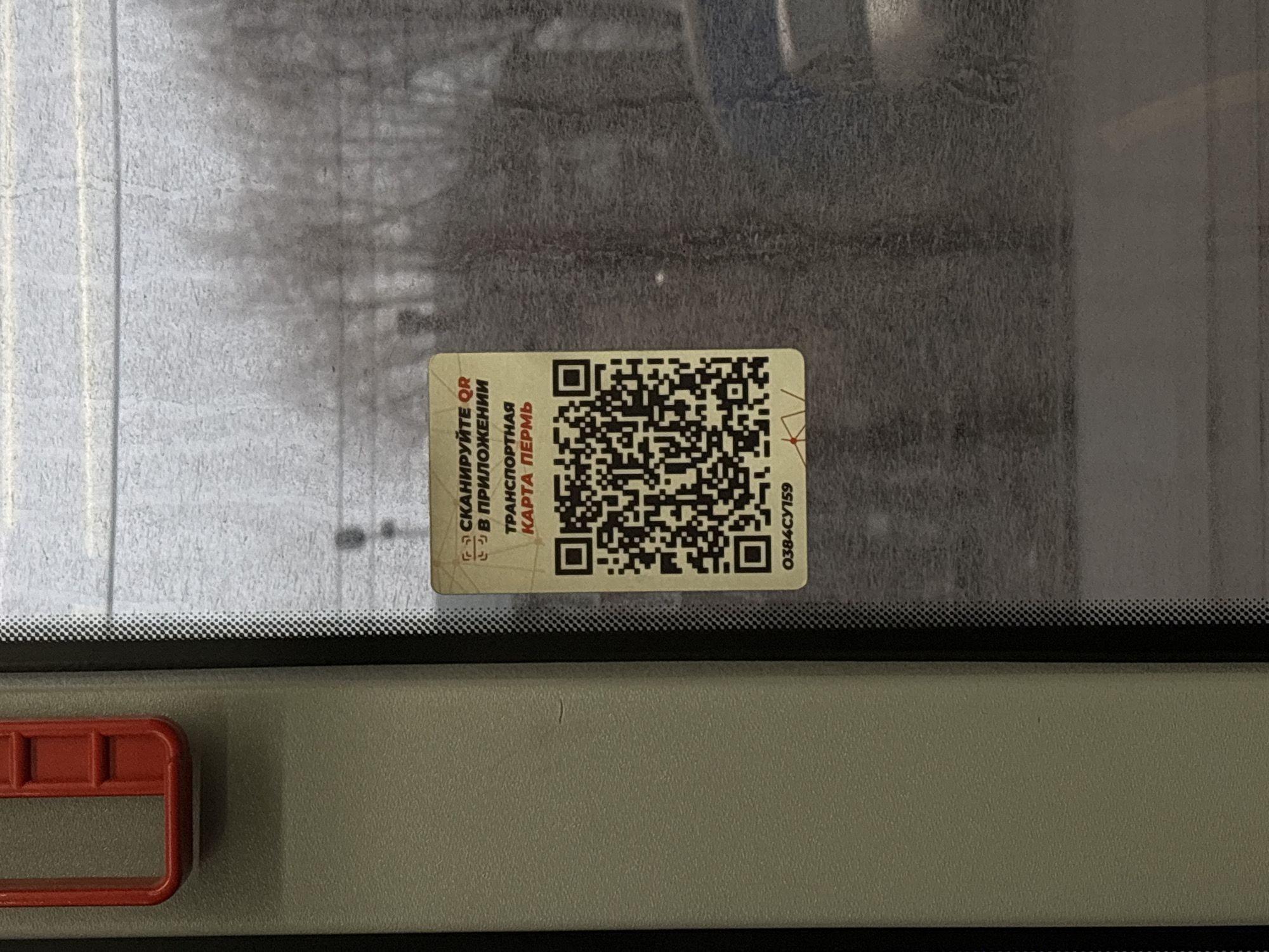 The image shows a QR code sticker on the inside of a vehicle window, likely a bus or tram. The sticker reads "Сканируйте QR в приложении Транспортная карта ПЕРМЬ", which translates to "Scan the QR code in the Transport card PERM application". The presence of the name "ПЕРМЬ" suggests the city of Perm in Russia as the location. Below the QR code, the alphanumeric string "0384CY159" is visible. The area around the sticker is out of focus, showing an indistinct view through the window, and part of an interior rail, possibly for standing passengers, is visible.