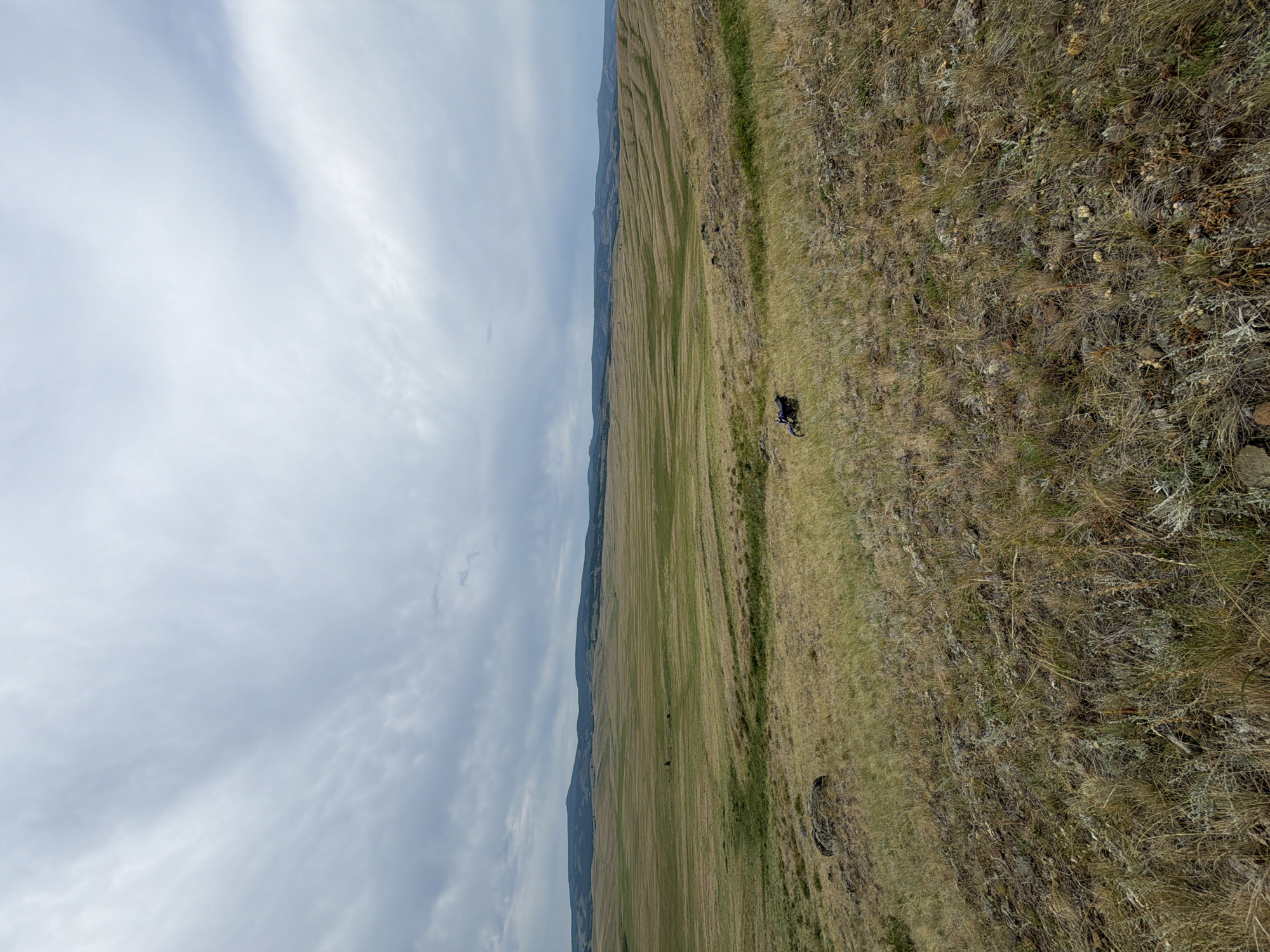 The image depicts a vast, undulating grassland under a cloudy sky. The terrain is characterized by rolling hills covered in dry, yellowish-brown grass, interspersed with patches of greener vegetation. In the middle ground, a dark-colored motorcycle is parked on the grassy slope. The background features a range of low, hazy mountains and hills stretching across the horizon. The foreground is rocky and covered with sparse, dry vegetation. The overall impression is one of a remote, natural landscape. The location is identified as Kuseevo, Russia.