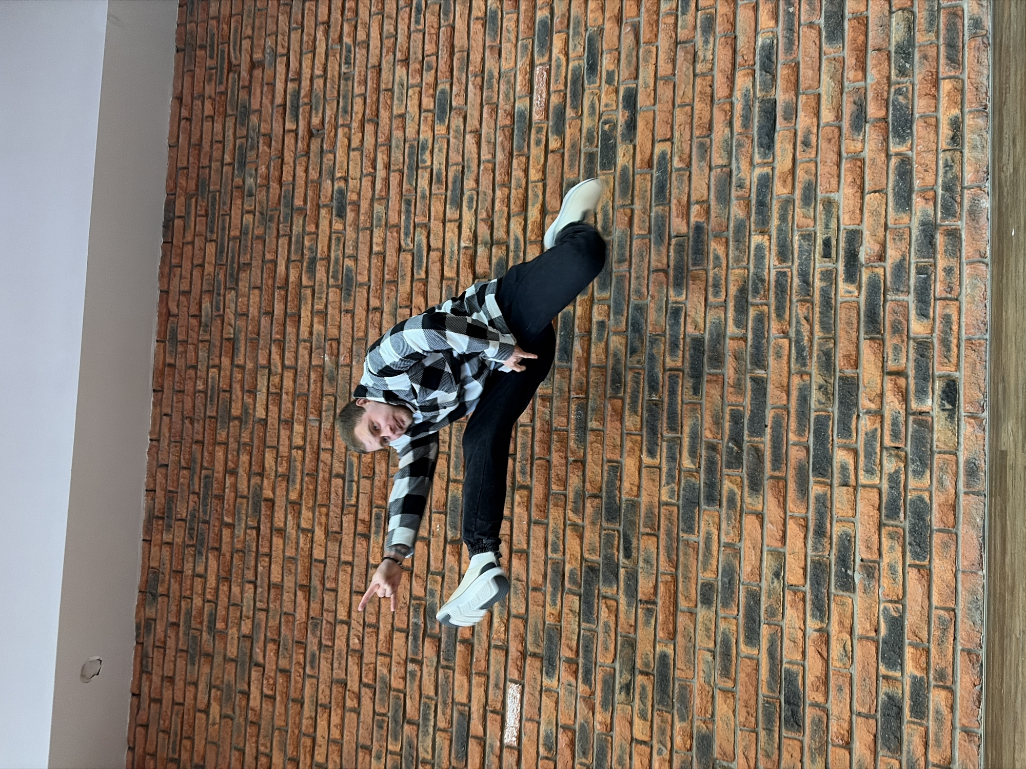 A man wearing a black and white checkered shirt and black pants poses indoors against a brick wall. He points with his left hand and bends his left knee, while his right leg is extended and slightly bent. The wall is a mix of red, brown, and black bricks and is joined by a white wall on the left and a wooden panel on the right. A small circular indent is visible on the white wall. The image was taken with the subject sideways, but it is not possible to determine the location in Chaikovsky, Russia without additional information.