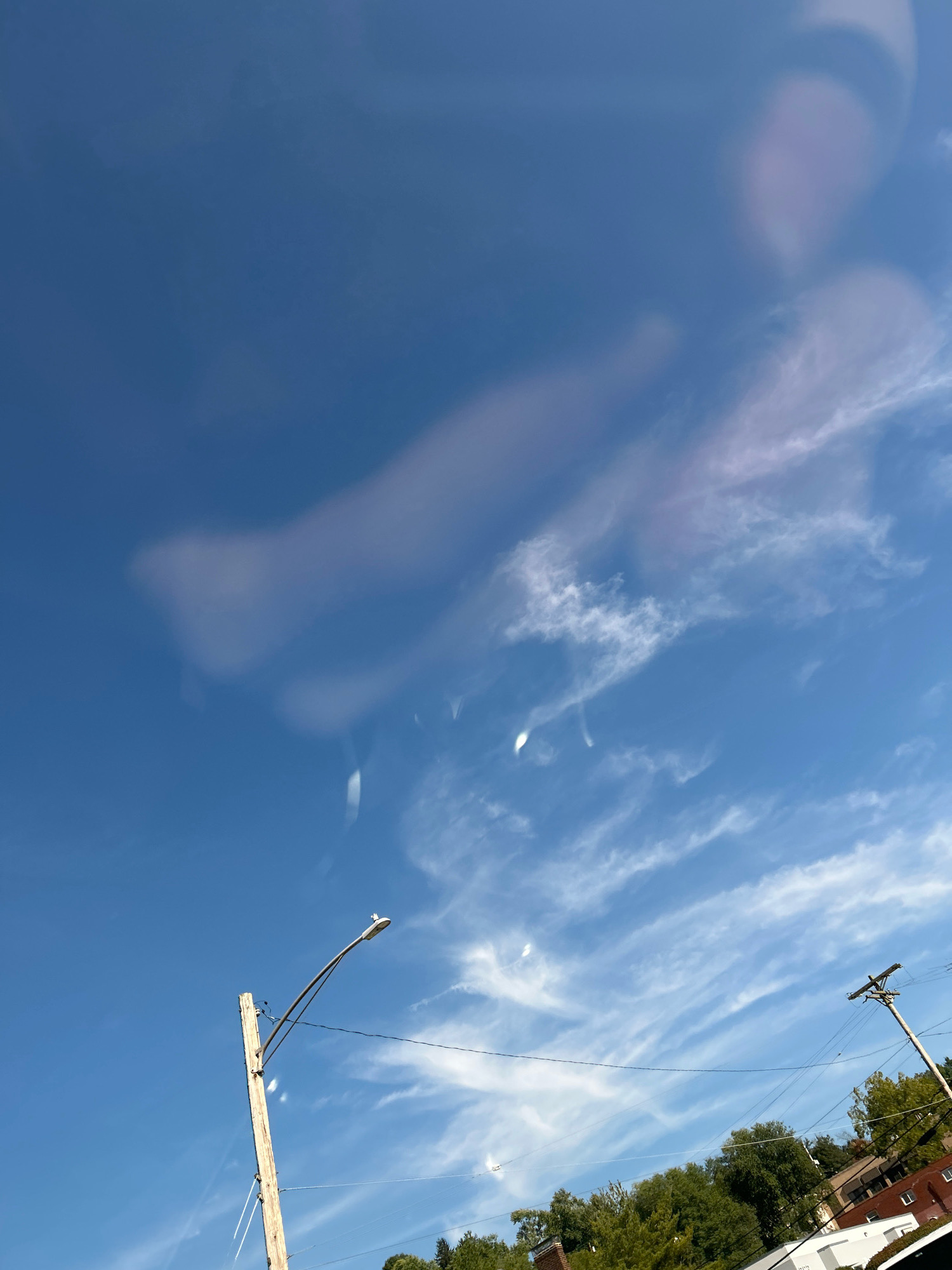 The image captures a bright blue sky with wispy, elongated clouds. In the foreground, a weathered wooden utility pole stands vertically, supporting a street light with a curved arm. Electrical wires stretch horizontally across the frame, connecting to another utility pole on the right side of the image. Beyond the poles and wires, a line of lush green trees and parts of buildings are visible, suggesting an urban or suburban setting. The overall impression is one of a clear day with scattered cloud formations. The scene is set in Pittsburgh, United States of America.