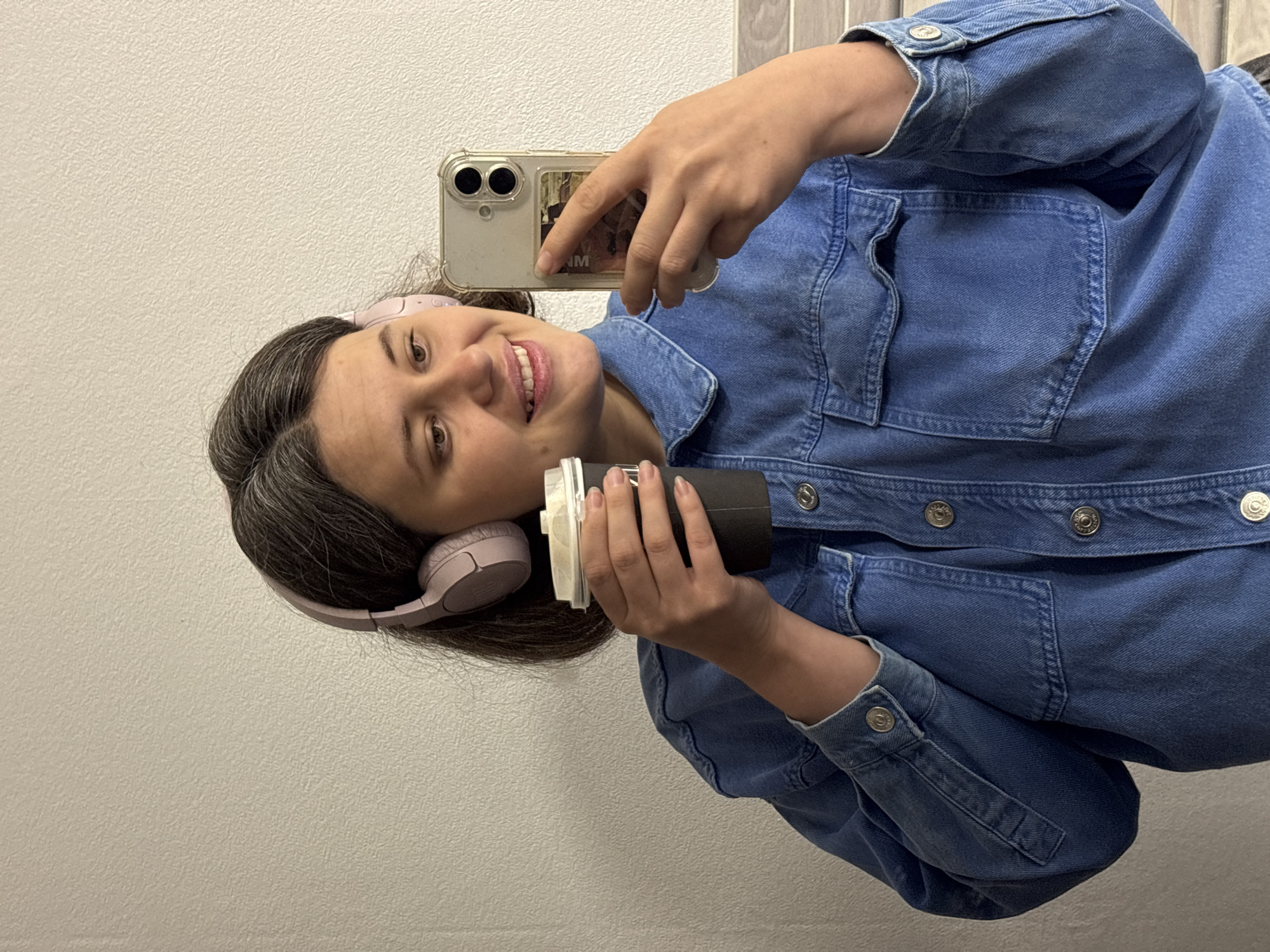 A young woman with short, dark hair and light-colored headphones is holding a disposable coffee cup in her left hand and a smartphone in her right hand, taking a selfie in front of a white, textured wall. She is wearing a blue denim jacket. The background appears to be an indoor setting with a tiled wall visible to the right. There is no visible indication of the city of Chaikovsky, Russia.