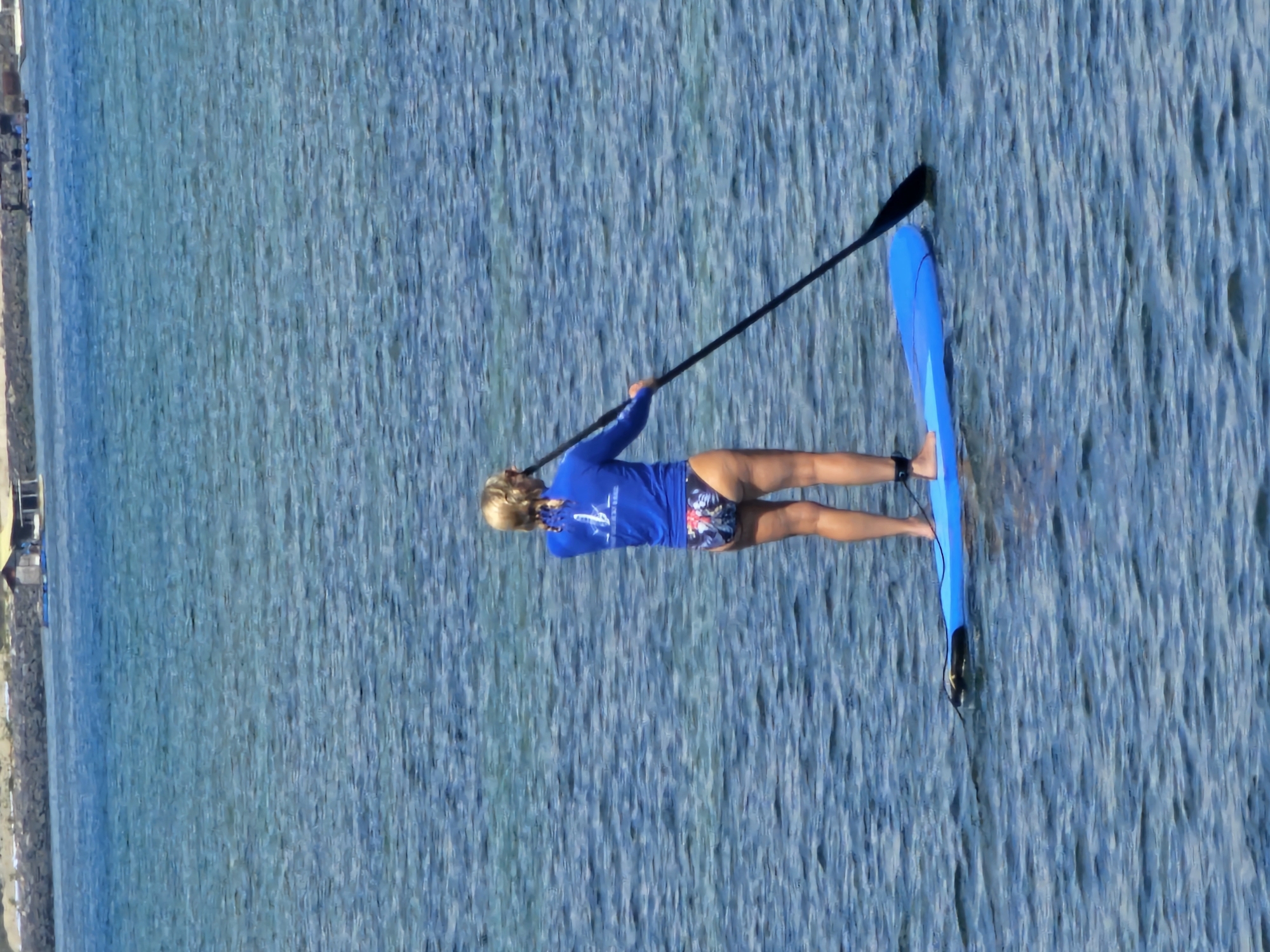 A woman with blonde hair, wearing a blue rash guard and floral print bikini bottoms, paddleboards on a bright blue board in the ocean. She is facing away from the camera, holding a black paddle. The water is calm with slight ripples, and the seabed is visible in some areas. In the background, a rocky shoreline and a small building on stilts are visible. The scene appears to be a recreational activity on a sunny day.