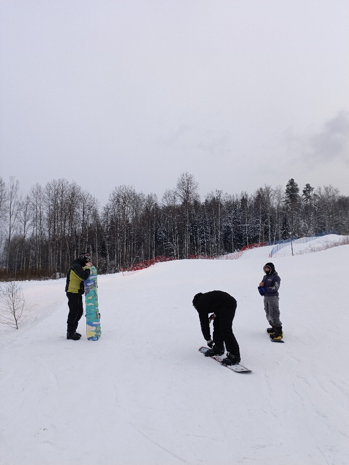 The image captures a winter scene on a snow-covered slope, likely a ski resort or snowboarding park, under an overcast sky. Three individuals are present on the white expanse, engaged with their snowboards.
In the foreground to the left, a person is standing upright, holding a snowboard vertically in front of them. This individual is dressed in a vibrant lime green and black jacket, black pants, and appears to be wearing a dark helmet or balaclava. The snowboard itself features a distinctive camouflage-like pattern in shades of light blue, yellow, and green.
Slightly to the right and in the mid-ground, another person is bent over, facing away from the viewer. This individual is entirely clad in black attire, including a jacket and pants, and is focused on adjusting the bindings of their snowboard, which is lying flat on the snow beneath them.
Further to the right and slightly behind the person in black, a third individual stands upright on their snowboard. This person is looking towards the left side of the frame and is wearing a grey hooded jacket, grey pants, and a dark helmet or goggles. Their snowboard is primarily yellow with black accents.
The background features a dense forest of trees, many of which are deciduous and bare, though some evergreen trees are also visible, all lightly dusted with snow. Running along the edge of the slope, behind the people, are safety nets or fencing, distinguished by sections of red and blue, indicating boundaries or hazard areas. The sky is a uniform, pale grey, suggesting a cloudy winter day without direct sunlight. There is no visible text in the image. The overall impression is one of a recreational winter outing, with the individuals preparing for or taking a break from snowboarding.