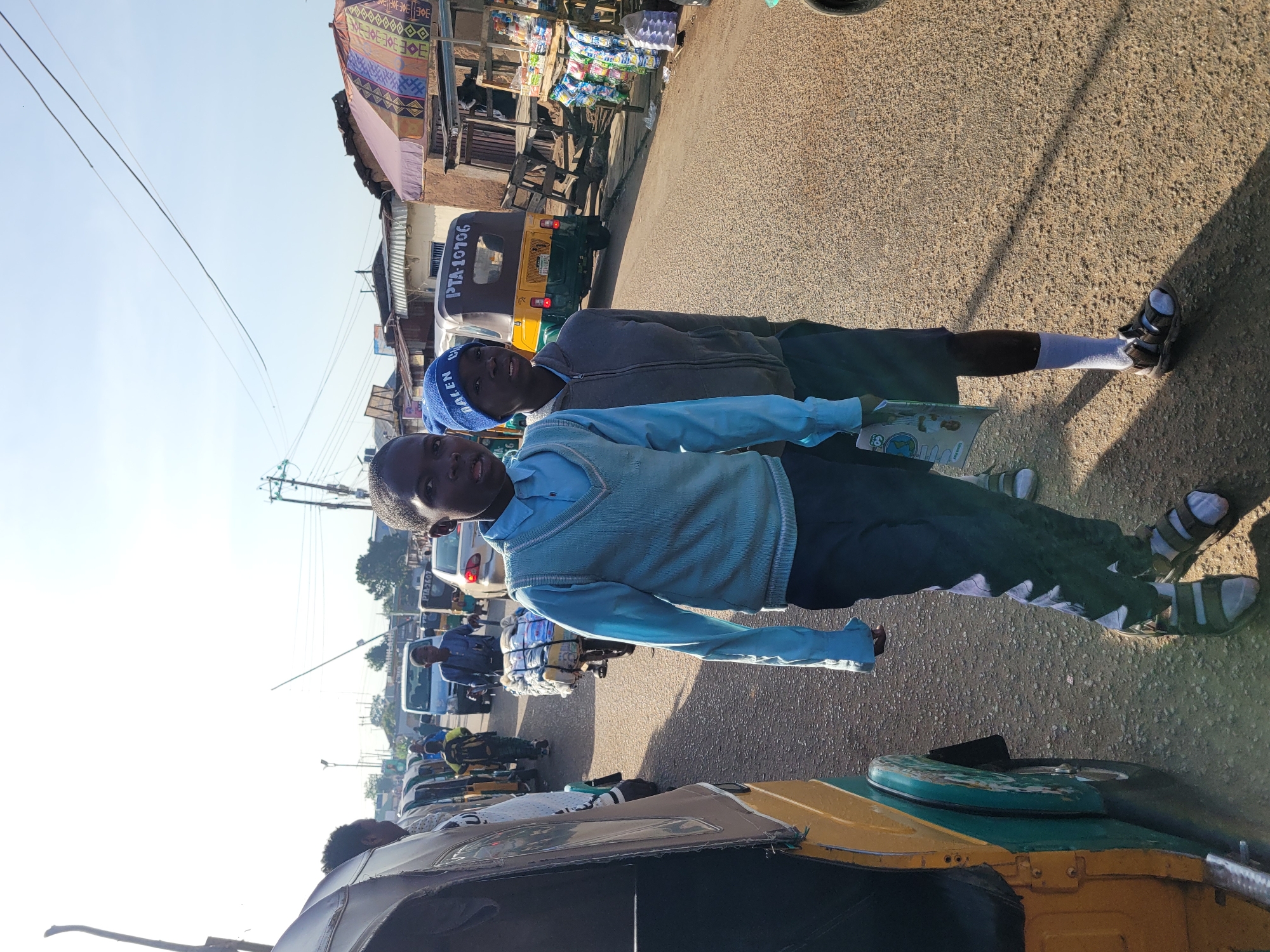 Two young boys stand on a dusty road in Jos, Nigeria. The boy on the left wears a light blue v-neck sweater over a long-sleeved shirt and dark pants, with sandals and socks. He holds a book in his left hand. The boy on the right wears a grey hooded jacket over a t-shirt and dark shorts, with sandals and socks. He has a blue knit cap with the word "DALEN" on it.
They are standing near a row of parked auto-rickshaws, some of which are yellow and green. Behind them, the road is lined with small shops and stalls displaying goods, and a few other auto-rickshaws and a car are visible. The sun is bright, casting shadows on the road, and utility poles with wires are present in the background. The scene appears to be a busy market or street in a residential area.