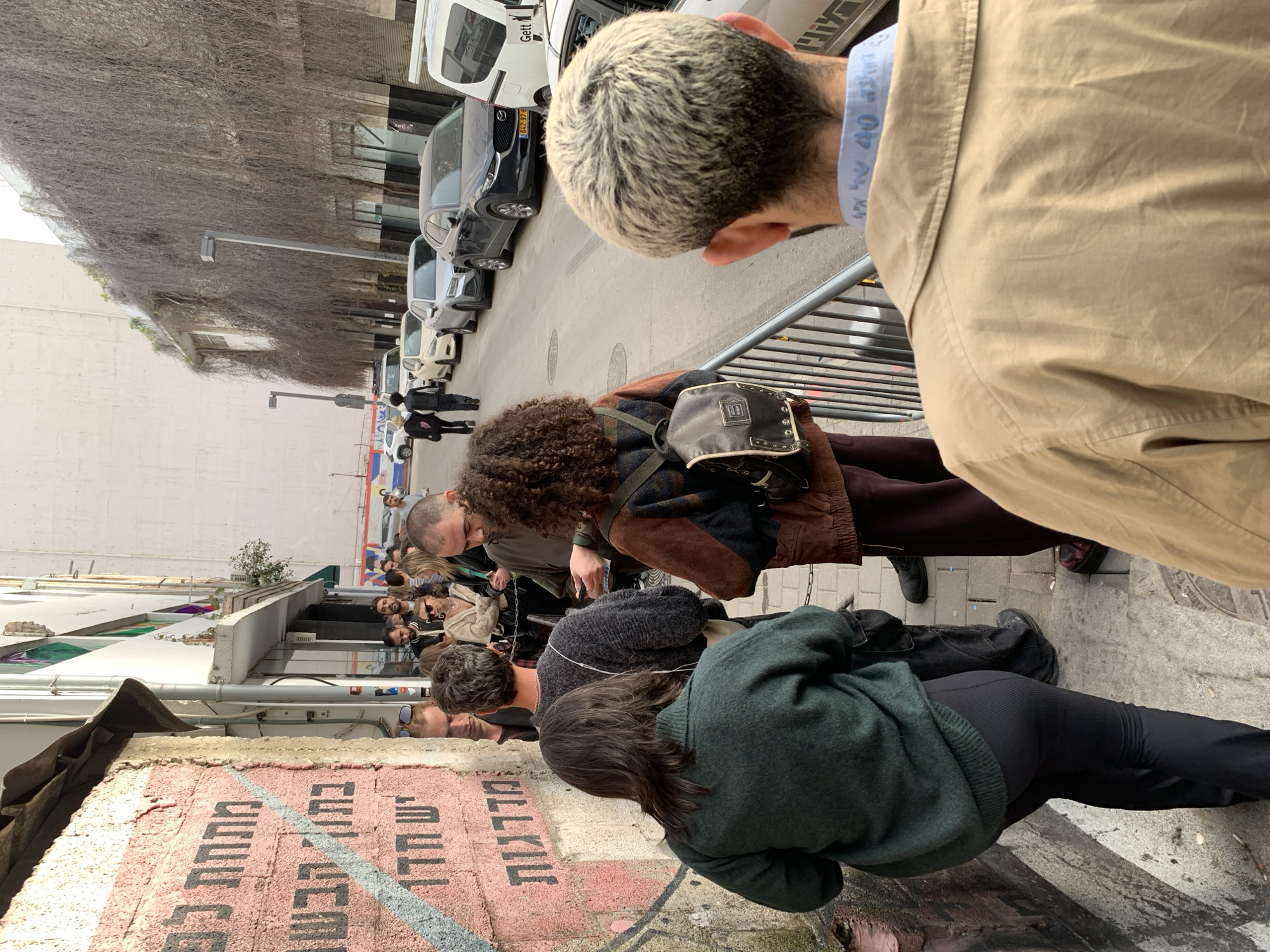 A horizontal eye-level photograph captures a street scene in Tel Aviv, Israel. A crowd of people is queued against a graffiti-covered wall, their bodies facing away from the viewer. The wall has faded, coral-colored paint, overlaid with black Hebrew lettering and a faded blue diagonal line. The clothing of the people in the queue is varied, with many wearing sweaters and coats. A man with bleached hair stands in the foreground, turned away from the camera. Metal railings run parallel to the queue. Beyond the railings, parked cars line the street, and a vine-covered building rises in the background. The sky is overcast.