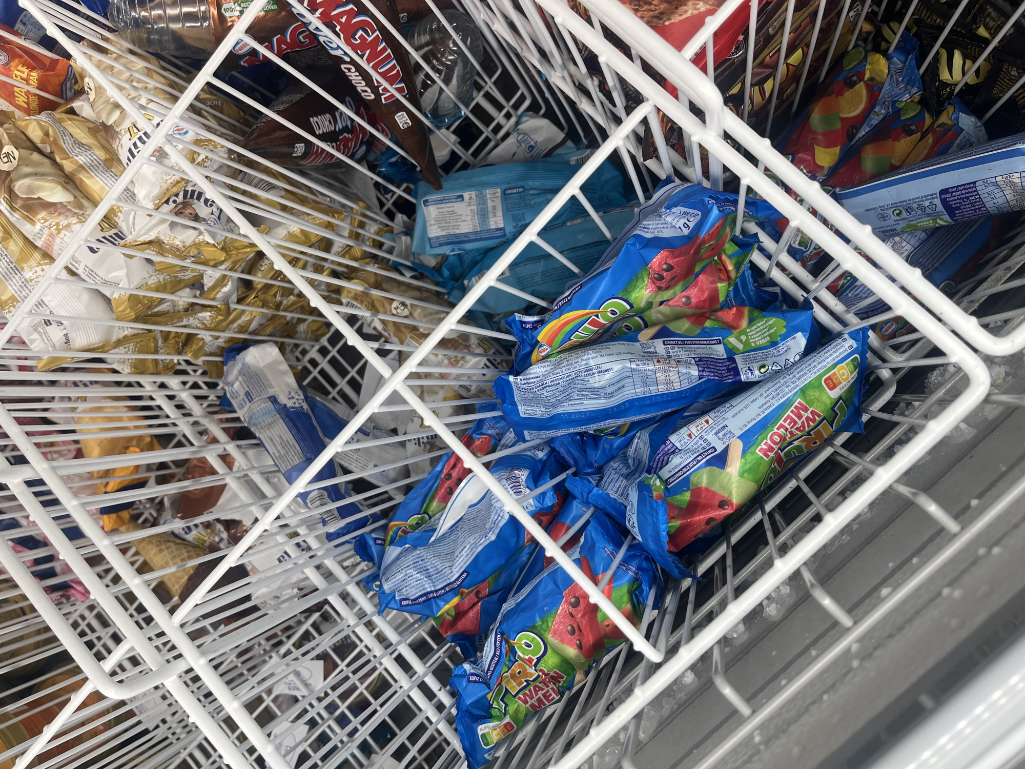 The image shows a freezer bin filled with various ice cream products. Several packages of ice cream are visible:
* "Pirulo Watermelon" popsicles are in the foreground, packaged in blue wrappers with images of watermelons.
* "Magnum" ice cream bars, identified by their distinctive packaging.
* Other ice cream types, some in gold-colored wrappers.
The bin is constructed from white-coated metal wire. The interior of the freezer appears metallic. Text on the wrappers mentions "София, 1220, България" which identifies the image location as Sofia, Bulgaria. The wrappers also include nutritional information and company contact details in multiple languages.