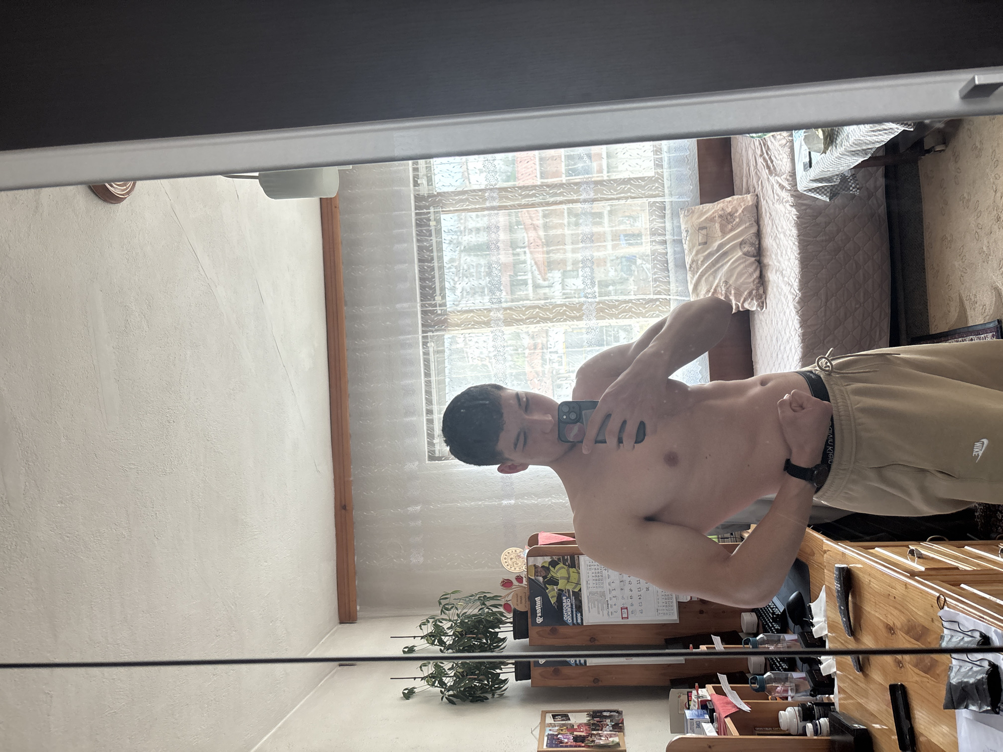 A shirtless young man stands in front of a mirror, flexing his arm and holding up a smartphone, presumably to take a photo of himself. He is wearing beige sweatpants. The reflection in the mirror shows a room with white walls, a window with a patterned curtain, and a bed. Below the mirror is a wooden shelf holding various items, including plants, a calendar, and miscellaneous objects. It is not possible to confirm the location as the city of Zlatograd, Bulgaria.