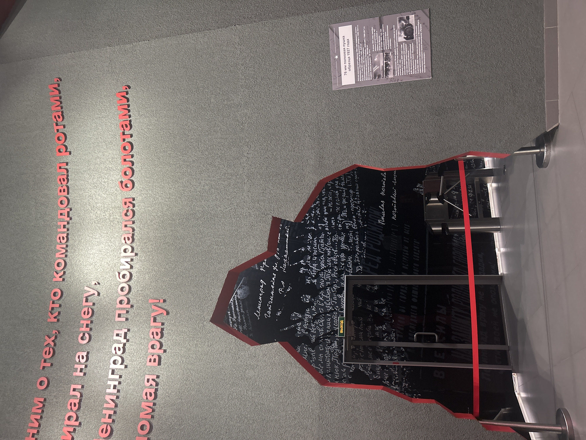 The image depicts a section of a museum exhibit. Dominating the left side is a large, irregularly shaped cutout in the wall, resembling a dark, jagged opening. This cutout is filled with a black surface upon which handwritten text is displayed in white, appearing to be excerpts from historical documents or personal accounts. Red lettering is affixed to the grey textured wall surrounding the cutout, forming Russian text that appears to be a quote. To the right, a portion of a grey wall is visible, and mounted on it is a white informational placard with Russian text and images, likely describing an artifact or historical event. The placard mentions "76-мм полковая пушка образца 1927 года" (76-mm regimental gun model 1927). A red velvet rope barrier is visible in the foreground, stretching horizontally across the lower part of the frame, indicating a boundary for visitors. The overall scene suggests an exhibition focused on military history, possibly related to the Siege of Leningrad, given the text on the wall and the presence of historical artifacts. The location is identified as Kirovsk, Russia.
