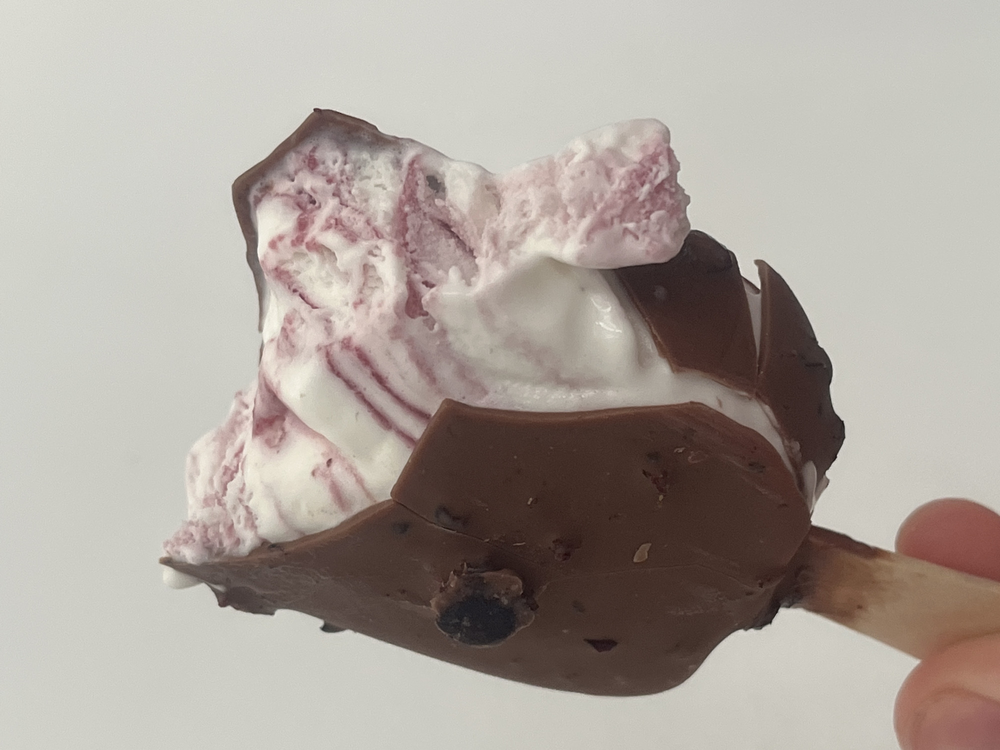 The provided image depicts a partially consumed chocolate-covered ice cream bar held by human fingers against a plain, light-colored background. No visual information within the image allows for the determination of a specific location in the city of Saint Petersburg, Russia, or any other geographical location.
The ice cream bar features a dark brown chocolate coating that has cracked and peeled back, revealing the interior. The exposed ice cream is white with prominent swirls of a reddish-pink substance, suggesting a fruit or berry flavor. A significant portion of the ice cream on the upper right side is missing, indicating it has been bitten into. Small, dark, solid fragments, possibly inclusions like berry pieces or chocolate crumbs, are embedded in both the chocolate coating and the exposed ice cream. One distinct, dark, spherical inclusion is visible on the left side of the chocolate coating. The ice cream is attached to a light-brown wooden stick, which is partially obscured by the bottom portion of the chocolate coating. The stick is held between the thumb and forefinger of a person, with only the skin-toned fingers visible at the bottom left of the frame. The background is uniformly light grey or off-white and appears out of focus, providing no environmental context.