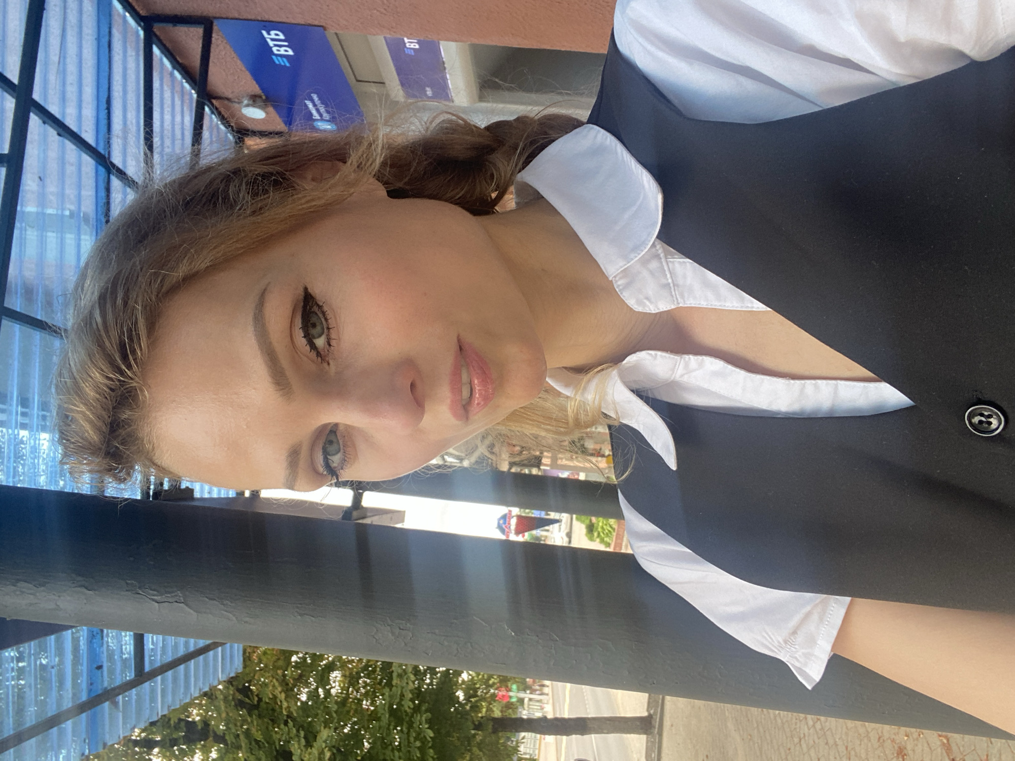 A young woman with blonde hair styled in a braid and subtle makeup is captured in a selfie taken outdoors. She is wearing a white collared shirt underneath a black vest with a single button closure. The background reveals a street scene in the city of Lida, Belarus, with a prominent building displaying the "ВТБ" (VTB) logo, indicating a bank or financial institution. A bus stop shelter with a transparent roof and a dark, textured pillar are also visible, suggesting the selfie was taken at a public transport stop. Trees and a street with vehicles and pedestrians are discernible in the distance, hinting at a bright, daytime setting.