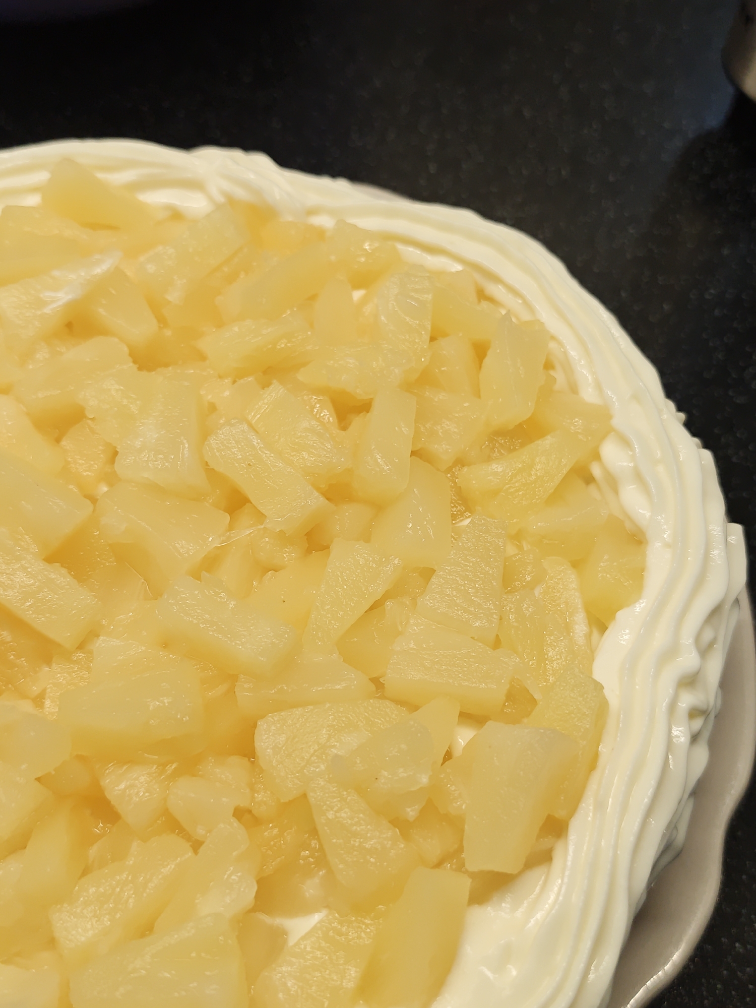 A close-up shot presents a cake with a creamy white frosting border and a topping of diced pineapple. The cake is on a light-colored plate, and the background is a dark, possibly black, surface. The pineapple pieces appear to be glistening, suggesting they may be in a syrup. The frosting has a piped design along the edge of the cake. The location is Unknown, Unknown.