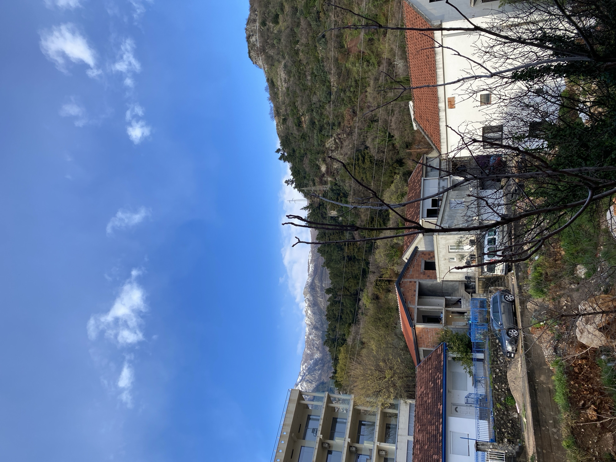 This image depicts a residential area within the city of Budva, Montenegro, characterized by a mountainous backdrop under a clear blue sky with scattered white clouds.
In the foreground, bare deciduous tree branches partially obscure the view of a sloping, uncultivated area with exposed soil and rocks. Immediately behind this, a narrow paved road is visible. A gray station wagon, possibly an Audi A4 or A6 Allroad, is parked facing right on the road. To its right, a white utility van is partially visible, also parked on the road.
The midground features several buildings. On the left, a single-story white house with a red tiled roof and blue metal railings on its porch is prominent. Adjacent to it, a two-story unfinished brick building with exposed concrete framing stands, showing signs of ongoing construction. To the right of the unfinished structure, another single-story white building with a red tiled roof is visible.
In the background, a verdant, tree-covered mountain rises steeply. Power lines and utility poles are visible traversing this hillside. In the far distance on the left, a range of snow-capped mountains is apparent, beneath a dense line of evergreen trees. No individuals or active interactions are observed within the frame.