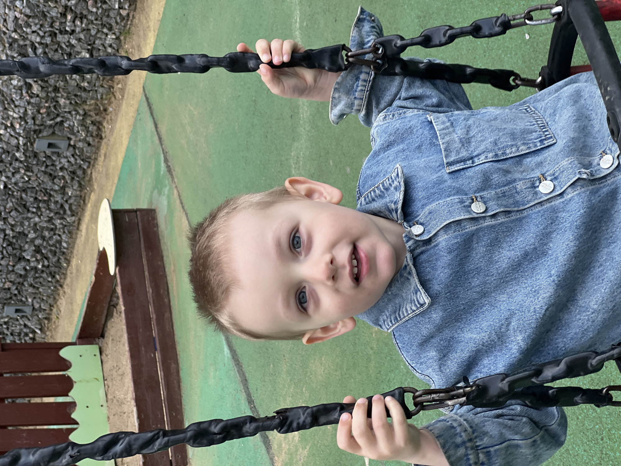 A young child with blond hair and blue eyes is holding onto the black, rubber-coated chains of playground equipment in Mytishchi, Russia. The child is wearing a light blue denim jacket with silver buttons. The background consists of green and brown play area surfaces. There is a wooden structure with a light green decorative trim visible in the background. The ground is covered in a mix of green turf and gravel.