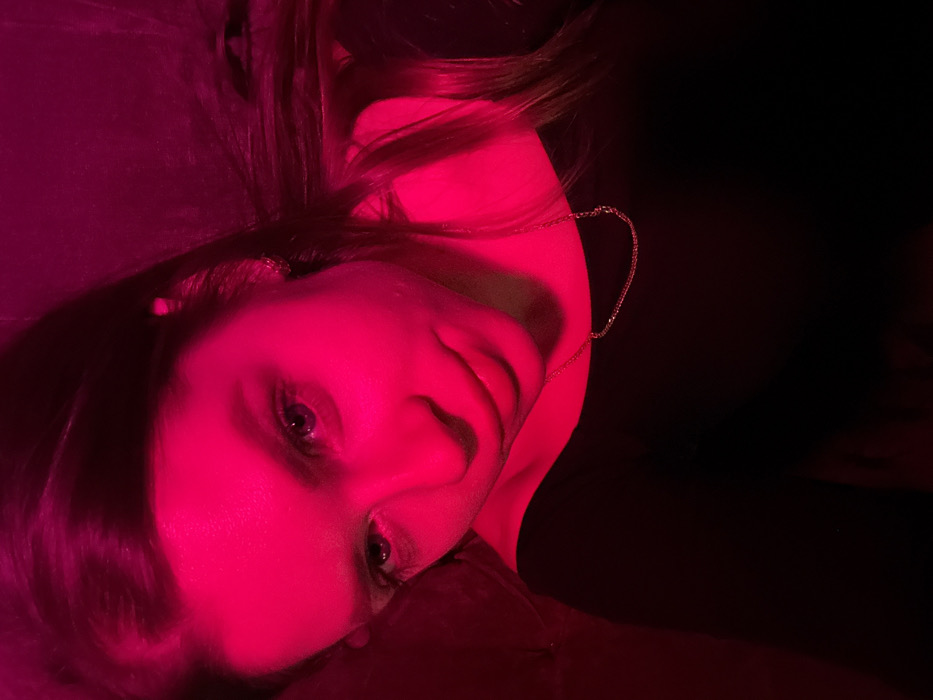 A woman's face is brightly lit in a hot pink hue against a stark black background. Her head is tilted, showcasing her eyes, nose, and lips under the intense magenta lighting. Part of her hair, also bathed in pink, frames her face. A delicate gold chain or earring is visible near her neck. The overall impression is a striking, almost surreal portrait due to the unconventional color palette.
