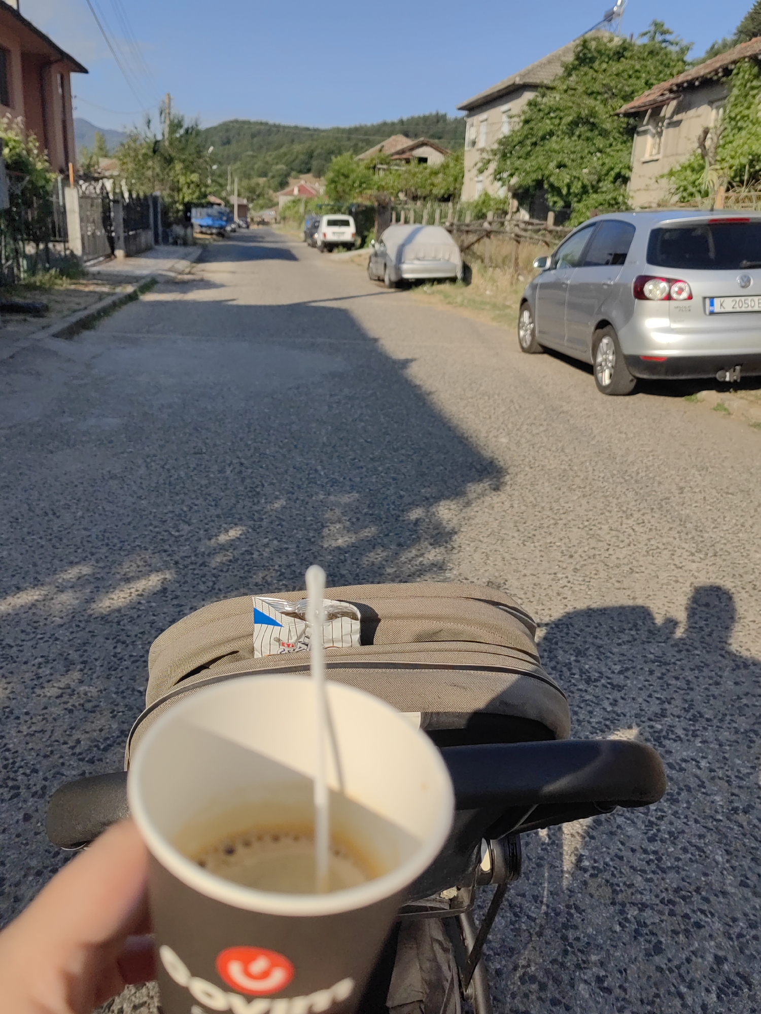 The image captures a scene on a street in the village of Djerovo, Bulgaria, bathed in the warm light of a sunny day. The perspective is from the point of view of someone on a bicycle, holding a disposable cup of coffee. In the foreground, a hand is visible holding the black paper cup with a red circular logo and white lettering. A white stirring stick is immersed in the coffee. Behind the cup, the back of a bicycle seat and a tan fabric bag are visible. The street is paved with asphalt, showing shadows cast by trees and perhaps the cyclist. Several cars are parked or driving along the street. A silver Volkswagen Golf is prominent on the right side of the road, with Bulgarian license plates. Further down the street, a white van and a blue truck are visible. Buildings with tiled roofs line the street, suggesting a residential area. Trees and vegetation are abundant, with a forested hill in the background. The sky is clear and blue.
