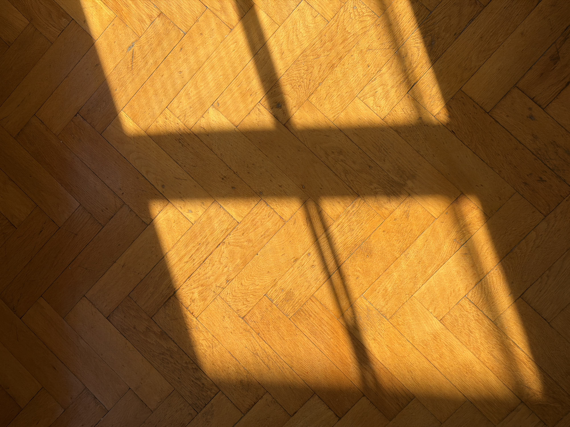 The image shows a close-up, overhead view of a wooden parquet floor with a herringbone pattern. Sunlight is casting shadows across the floor, creating a play of light and dark. The wood appears to be a warm, golden-brown color and shows signs of age and wear, with some scuffs and imperfections visible. The shadows suggest that the light source is a window, likely on a sunny day. The overall impression is one of a quiet, perhaps domestic, setting bathed in natural light. There is no visible text.
