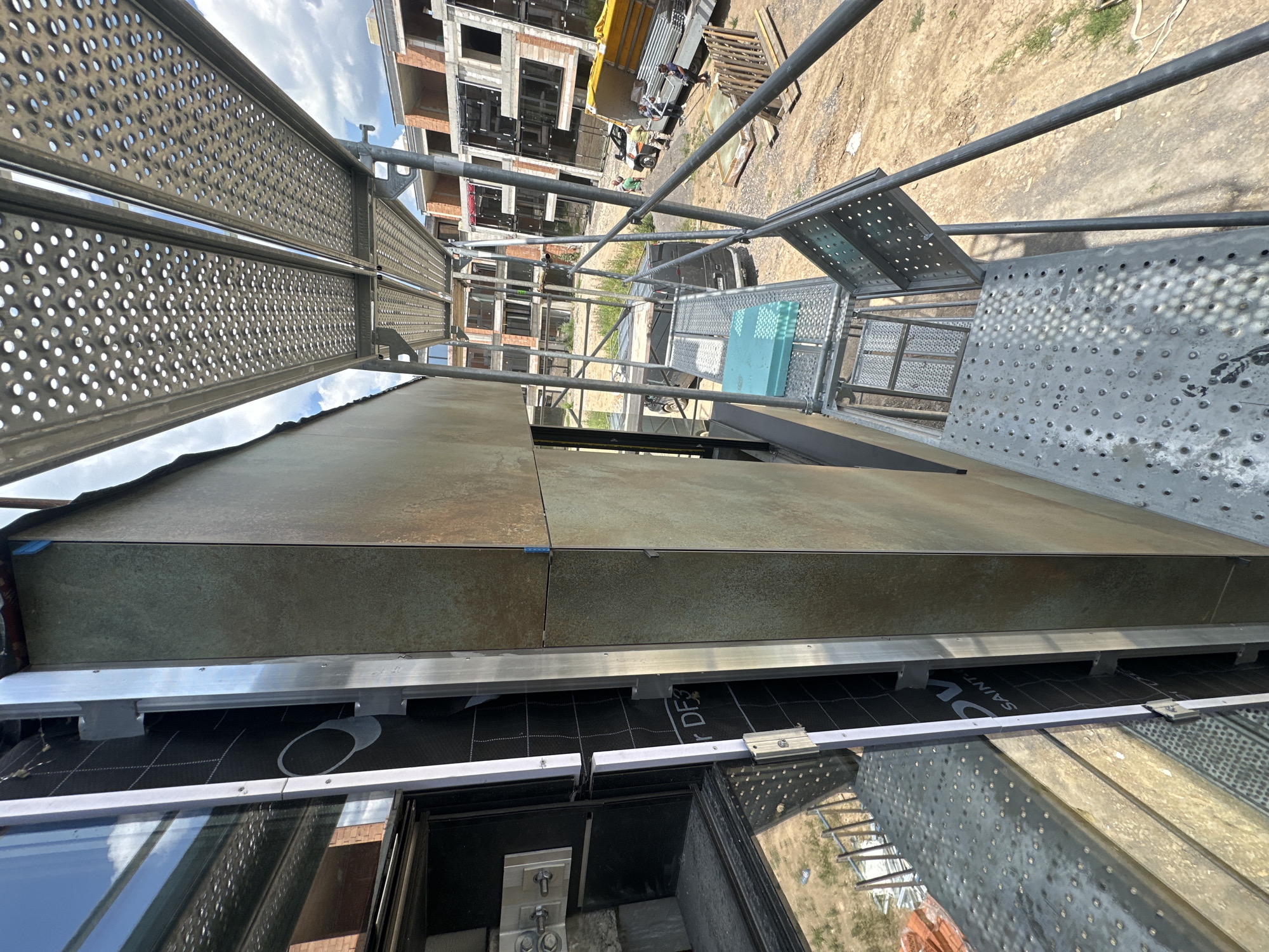 This media file captures a construction site in Stara Zagora, Bulgaria. The perspective is from a scaffolding platform, looking upwards and across a building under construction. The scene is bright and appears to be a sunny day.
The primary subjects are the scaffolding itself, made of metal poles and perforated metal planks, and the building facade. The building is in its early stages, with exposed brickwork, concrete structures, and unfinished window openings. In the background, other construction elements like a yellow truck, wooden pallets, and more building structures are visible, along with a dirt and gravel ground.
The activity taking place is construction work, evidenced by the scaffolding and the unfinished state of the building. While no people are clearly identifiable as distinct individuals in the foreground, the presence of construction materials and equipment suggests ongoing work. A few individuals can be seen in the mid-ground near a vehicle.
Notable details include the metallic texture of the scaffolding and the slightly weathered appearance of the building facade, which seems to be clad in large, textured tiles. The sky is partly cloudy with a bright blue hue. The image is oriented vertically, with the ground appearing in the upper portion of the frame.
Visible text on the black material beneath the facade reads "SAINT-GO..." and "...FDS", likely part of a brand name for construction materials.