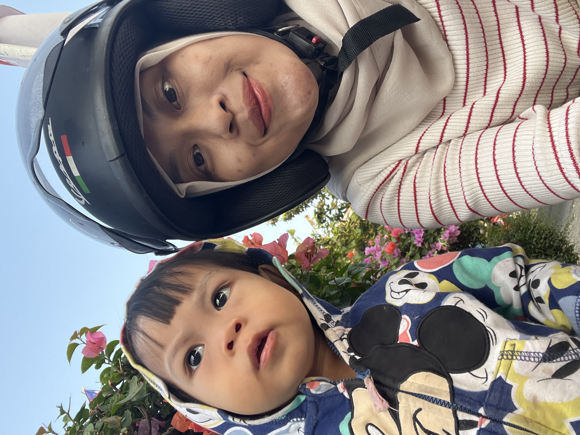 A close-up, eye-level, over-the-shoulder selfie shows two individuals on a street in Kecamatan Pati, Indonesia, with flowering bougainvillea in the background. On the left is a young child wearing a blue hooded jacket with a colorful Mickey Mouse print. The child is looking upwards and to the left. To the right is an adult, likely a woman, wearing a light-colored hijab and a white sweater with thin red stripes. She is wearing a black helmet with an Italian flag decal. The adult is smiling at the camera. The lighting suggests it is daytime, possibly late afternoon. The scene appears casual and informal.
