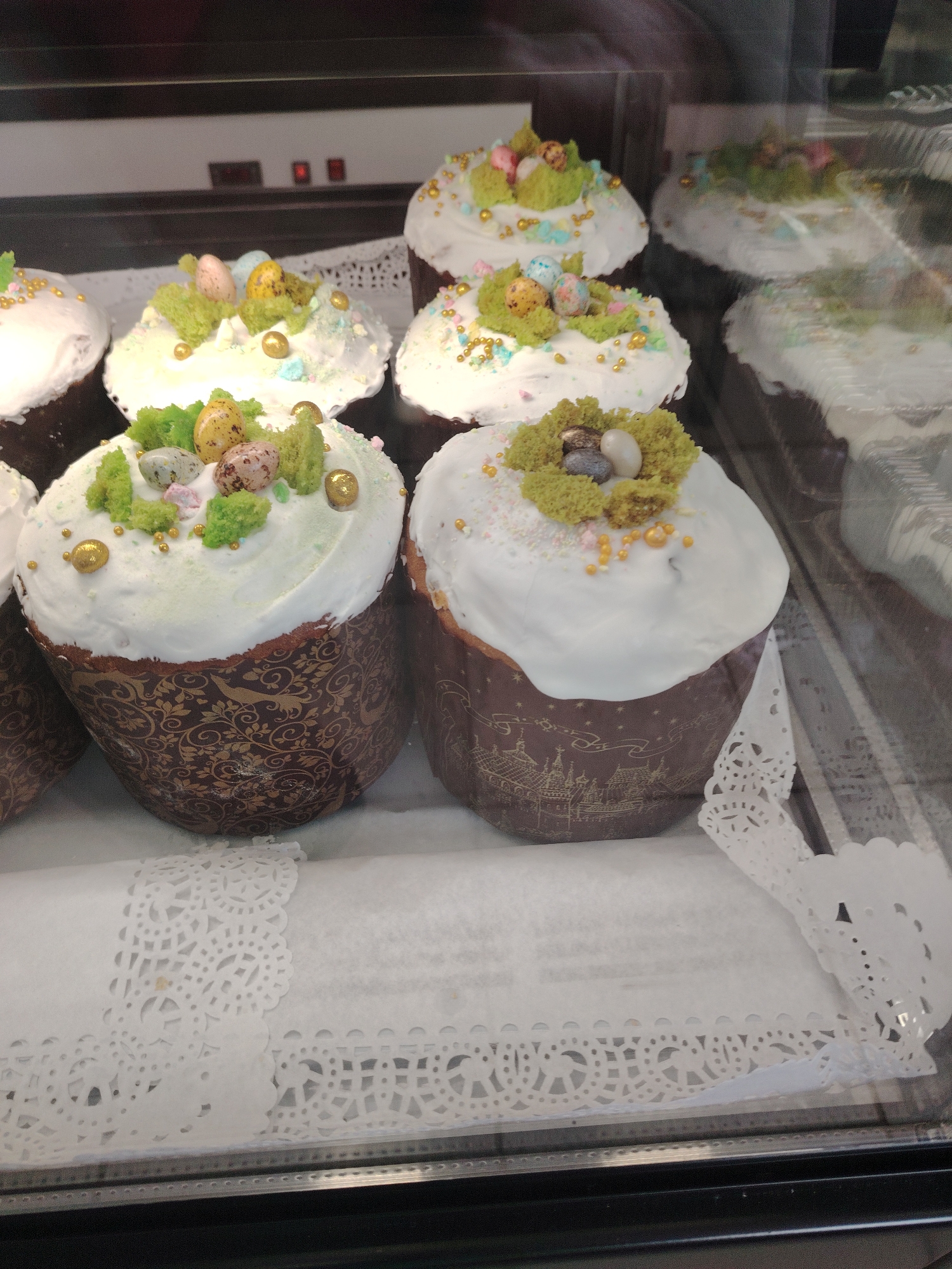 The image shows a display case filled with decorated Easter cakes, known as kulich, likely taken in Sarapul, Russia. The cakes have cylindrical paper wrappings with patterns; one shows a floral design and the other a depiction of buildings. Each cake is topped with white icing and decorative elements resembling nests with small eggs, green decorations resembling grass or foliage, and gold sprinkles. The cakes sit on a white, lacy paper doily inside the case, which appears to be part of a larger display in a bakery or shop. The reflection in the glass indicates the presence of other items in the display, potentially more kulich cakes. The focus is on the cakes in the foreground, with the cakes further back being somewhat blurred due to depth of field.