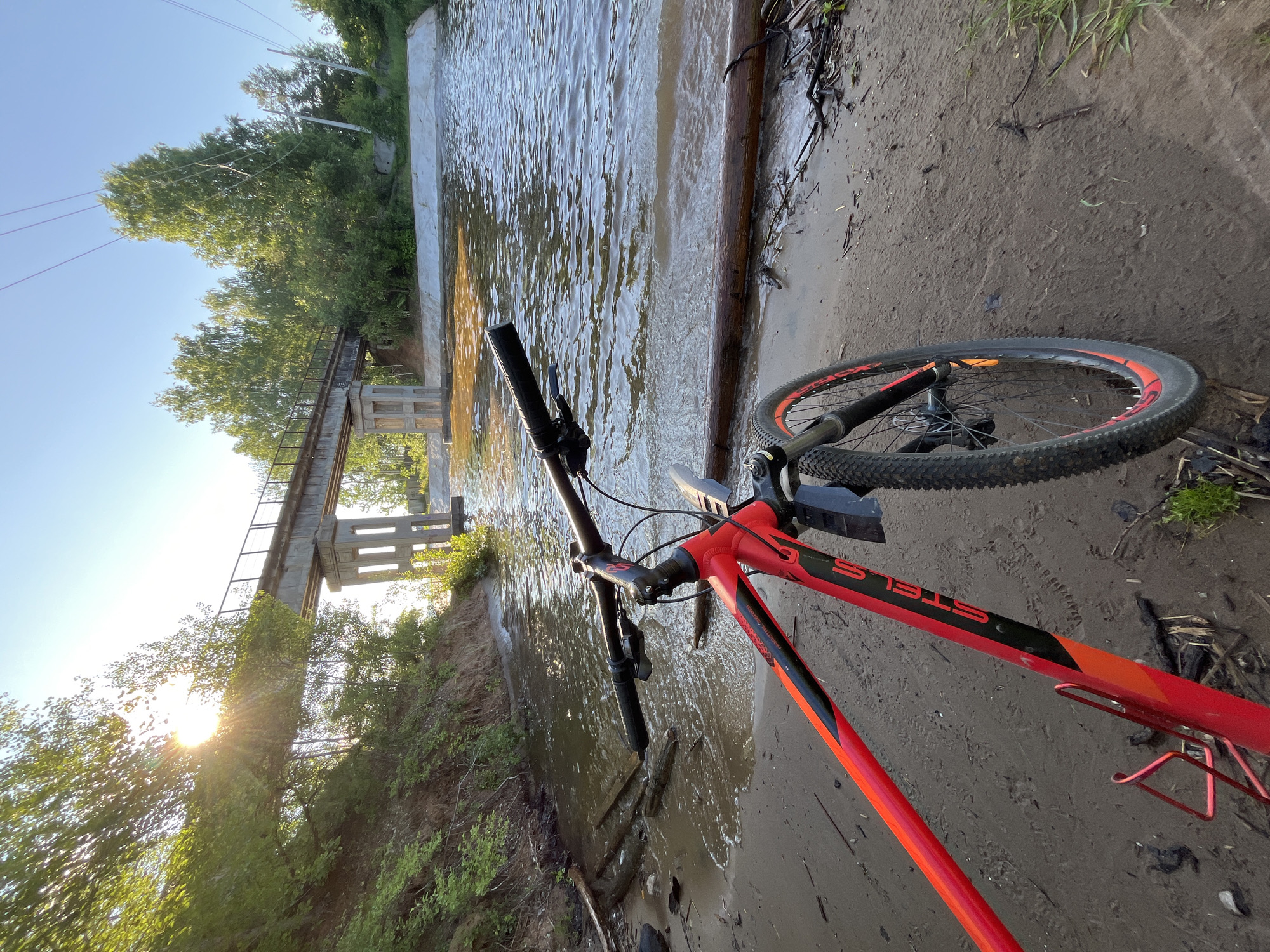 A red and black bicycle is parked on a sandy bank next to a body of water. The bicycle is in the foreground, with its front wheel turned towards the water. A concrete bridge with railings spans the water in the background, and trees line the opposite bank. The sun is setting, casting a warm glow on the scene. The location appears to be the Kama River in Chaikovsky, Russia, given the distinctive bridge structure and the general landscape. There are no people or animals visible in the image. The water is calm with some ripples, and debris such as logs and branches are scattered along the shoreline. Power lines are visible crossing the sky in the distance. The overall atmosphere is peaceful and serene.