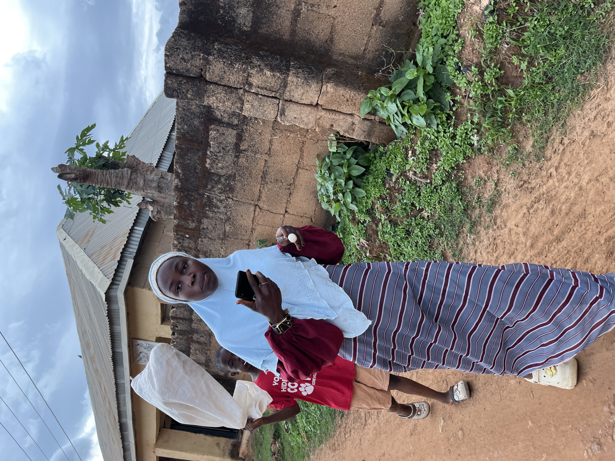 This image depicts an outdoor scene in what is described as the city of Jos, Nigeria, featuring two individuals on a reddish-brown dirt path.
In the foreground and mid-ground, a female adult is positioned slightly right of center, looking directly at the camera. She wears a light blue hijab with a white forehead band, a maroon long-sleeved top with voluminous sleeves, and a long striped skirt in shades of grey, blue, and maroon. Her footwear consists of light-colored closed-toe shoes with a yellow embellishment on the right shoe. She holds a black smartphone in her raised right hand and a small white spherical object in her left hand. A dark-colored bracelet with gold accents is visible on her right wrist.
Behind the woman and to her left, a young child is partially visible, walking in the same direction. The child wears a red t-shirt with white text and a paw print graphic, light brown shorts, and light-colored open-back clogs. The child carries a large, light-colored, possibly white, sack or bundle over their left shoulder.
The background features a weathered, dark-colored stone or mud-brick wall along the right side of the frame, showing vertical cracks and moss growth at its base. Green vegetation, including small leafy plants and grass, grows along the base of this wall and the dirt path. In the left background, a single-story building with light-colored walls and a corrugated metal roof is visible under an overcast sky with grey clouds. A pruned tree stump with green foliage sprouting from its top is located behind the wall in the right background. Electrical lines are faintly visible against the sky in the upper left quadrant. The ground is consistently reddish-brown dirt.