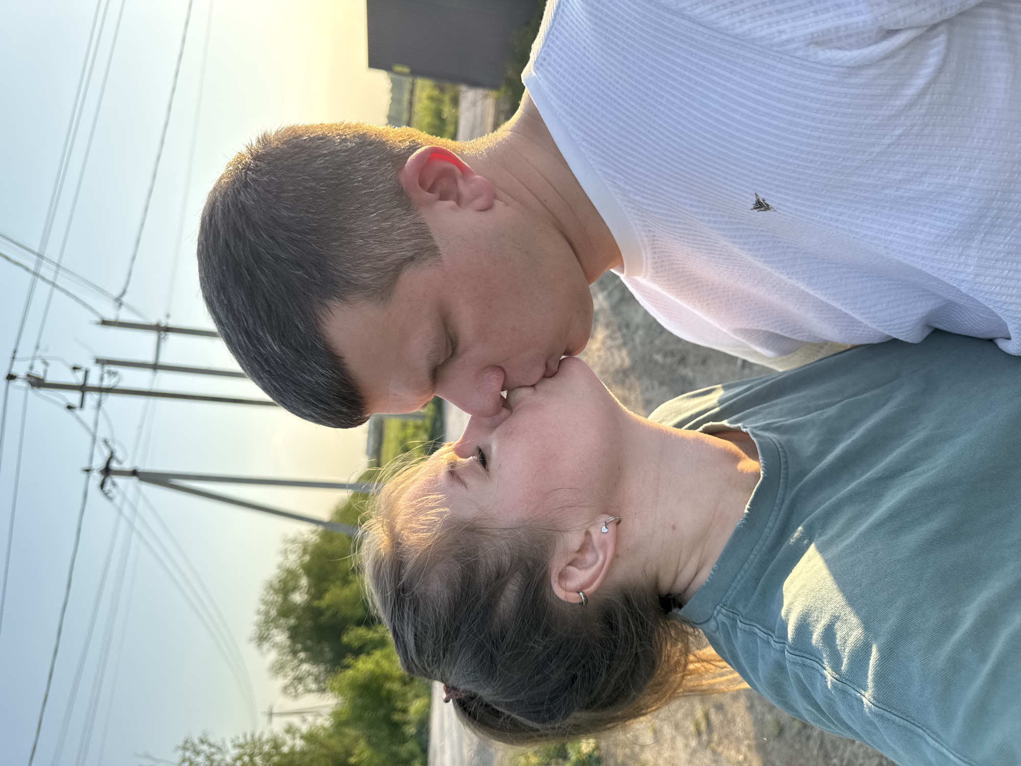 A couple is kissing in an outdoor setting near Горный, Russia. Power lines and vegetation are visible in the background. The man is wearing a white shirt, and the woman is wearing a green shirt. They are positioned close together.