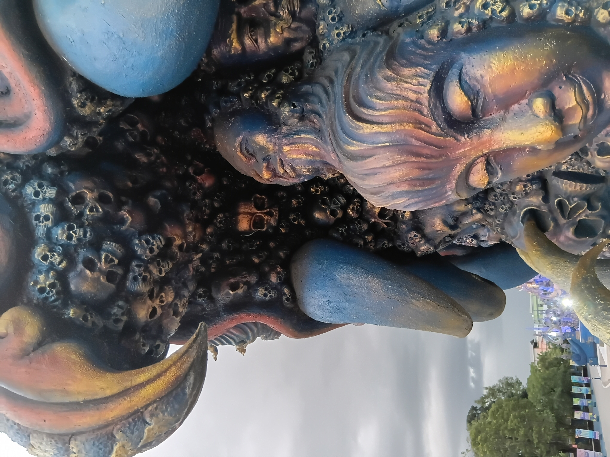 The image is a close-up of a detailed, multi-colored sculpture, likely a Buddhist temple or art installation, located outdoors in Chiang Rai City Municipality, Thailand. The sculpture is adorned with numerous human skulls, some appearing to be cast in relief and others more three-dimensional. Prominently featured are carved faces with serene, closed eyes, rendered in earthy tones with metallic highlights of gold and rust. The sculpture also includes large, bulbous shapes painted in a vibrant blue. In the background, a cloudy sky is visible above a row of trees and banners lining a pathway. Further in the distance, hints of a city skyline with illuminated structures suggest an urban setting. The overall impression is one of a surreal and intricate artistic creation, possibly depicting themes of life, death, and enlightenment, characteristic of religious art found in Thailand.