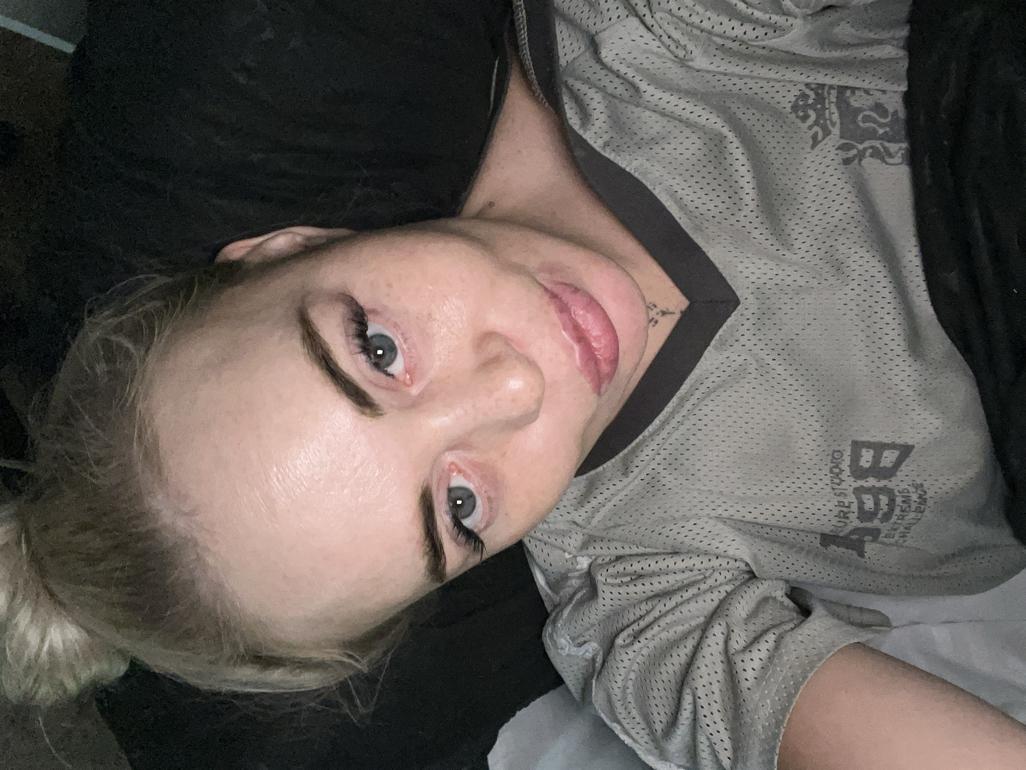 A close-up, overhead, selfie-style shot shows a white woman with blonde hair pulled up in a bun. She has blue eyes, fair skin with freckles, and is wearing a grey mesh athletic shirt. The shirt has a v-neck and a dark grey trim. There is a logo on the left side of the shirt that is partially obscured. A small tattoo of a cross is visible on her neck. The background is dark and out of focus, suggesting an indoor setting.