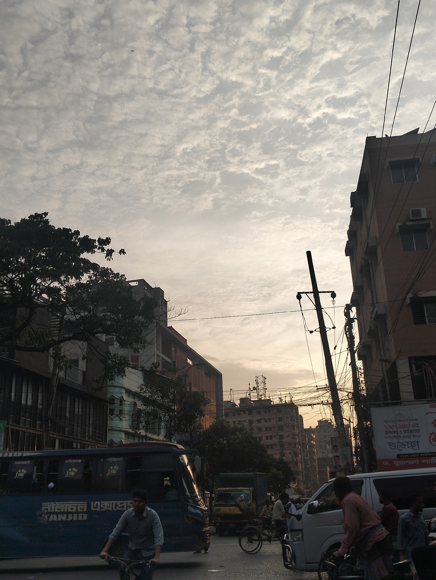 The image depicts a bustling street scene in Dhaka, Bangladesh. The sky is overcast with altocumulus clouds. A dark-colored bus labeled "নীলাচল এক্সপ্রেস Tanjid" is partially visible on the left, with a man riding a bicycle in front of it. Other vehicles, including vans and rickshaws, are seen amidst heavy traffic. Overhead, a dense network of utility wires crosses between buildings. Buildings of varying heights and architectural styles line the street. A vertical street sign is posted, promoting greeting for "Pabitra Eidul Fitar". There is an antenna/communications tower in the mid-ground.