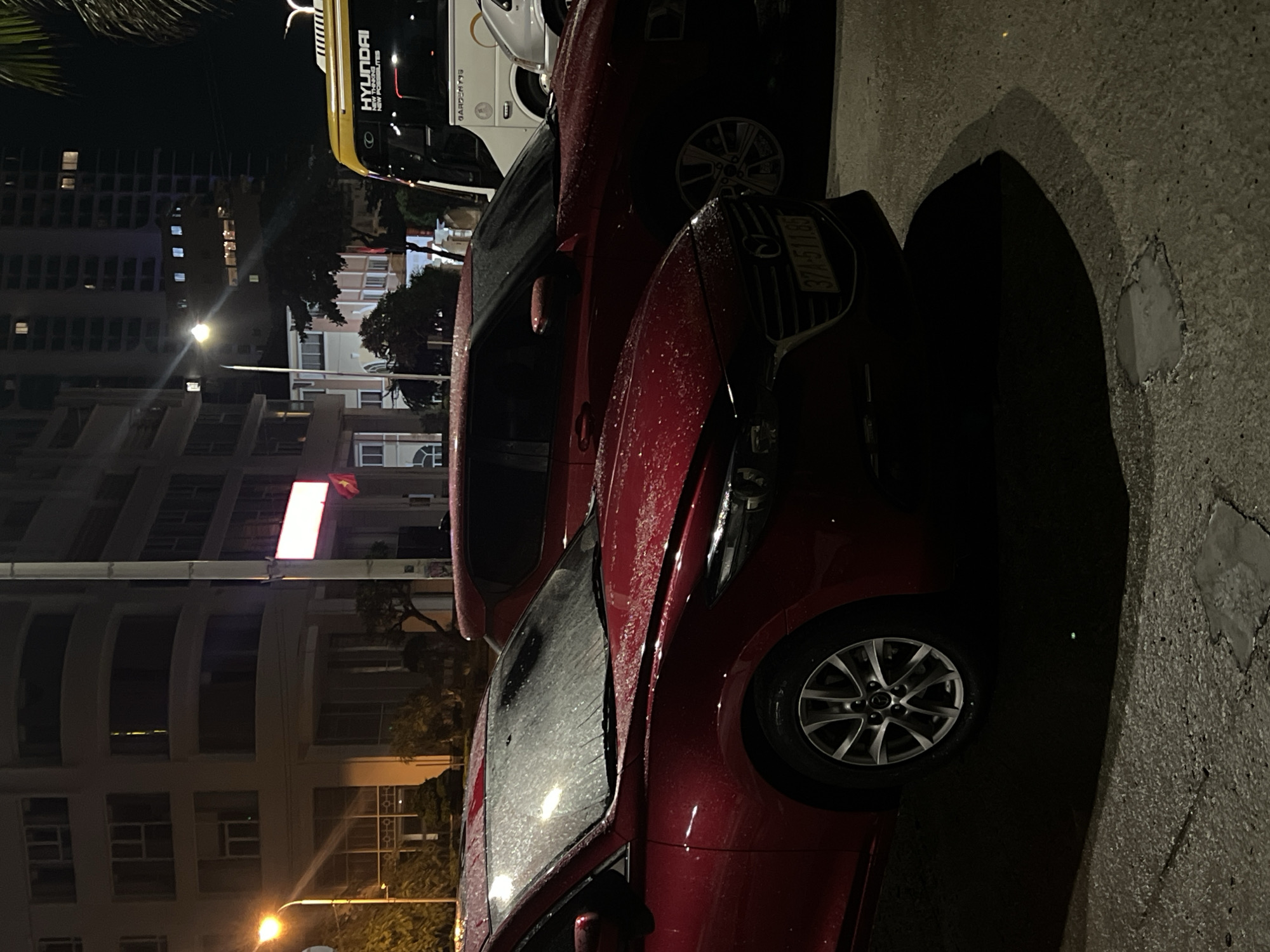 A rainy night in Bac Nha Trang Ward, Vietnam shows a red car parked on a wet street. A Hyundai vehicle is visible behind the car. Buildings line the street, illuminated by streetlights. The car's license plate is partially visible.