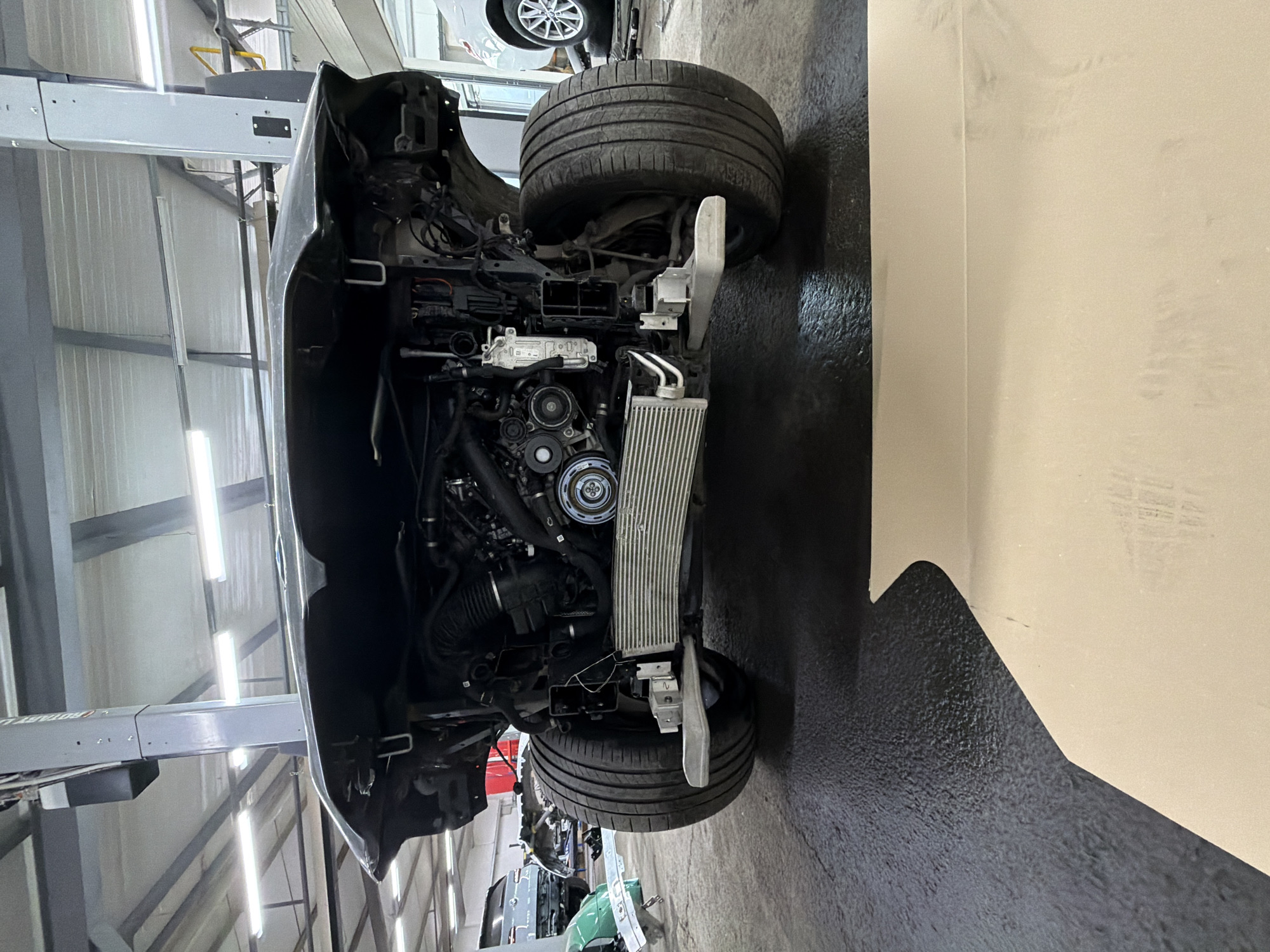 The image displays the front section of a car, partially disassembled, within an indoor automotive repair shop or garage. The car is oriented sideways in the photograph, showing its engine bay with various components visible, including parts of the engine, an air intake system, and a prominent silver radiator or intercooler unit positioned at the very front. A single front tire and wheel assembly are also clearly visible on the right side of the car.
The vehicle appears to be elevated on a hydraulic car lift, with a grey metal support structure visible on the left side of the frame. Fluorescent tube lights illuminate the workspace from above. The setting features light-colored, possibly corrugated metal walls in the background and a dark concrete floor. The overall impression is that of a professional service environment where the car is undergoing maintenance or repair.
Visible text includes the partial brand name "BODBOTT" on the grey car lift structure. Other smaller markings or labels on various car components are present but not legible. No people are visible in the frame, and the time of day is not discernible due to the indoor setting with artificial lighting.