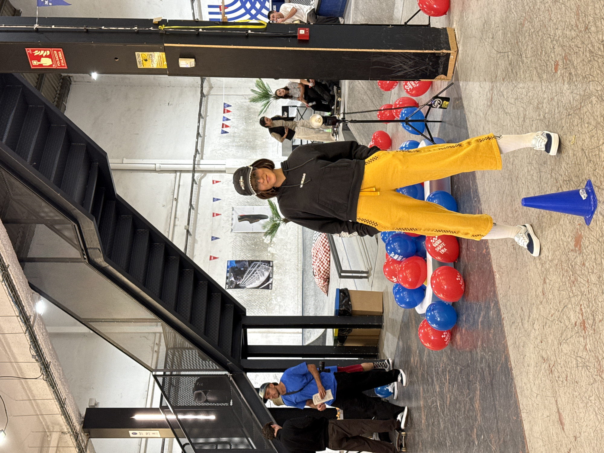A full shot shows a woman standing amidst red and blue balloons with "Vans Skate Era" printed on them. She wears a dark hoodie, yellow pants, and white socks with dark sneakers. A staircase with black metal railings is to the left. In the background, other people are present, some seated and observing. A blue cone sits near the woman. The location is L'Hospitalet De Llobregat, Spain.