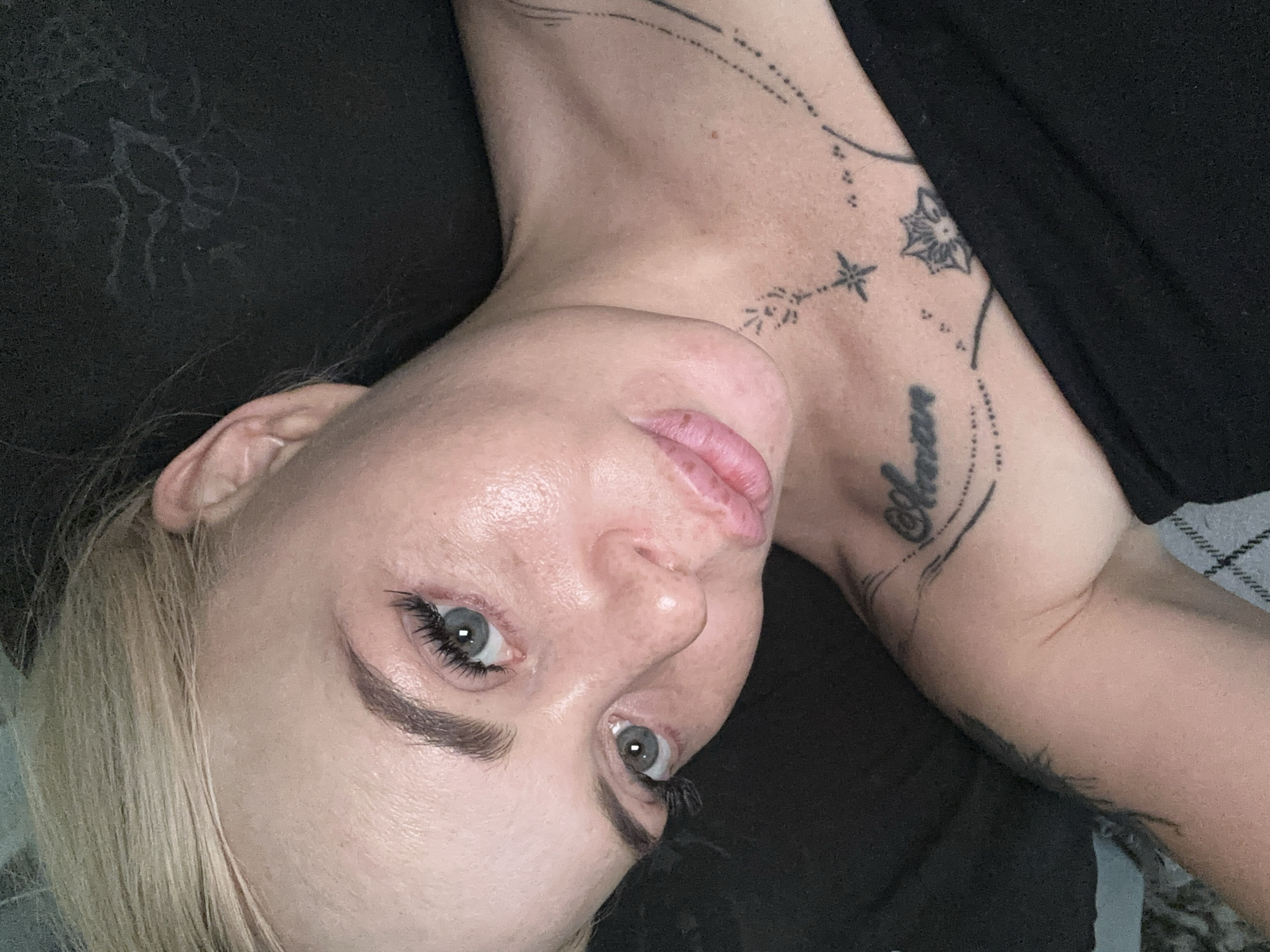 A close-up, high-angle, overhead shot focuses on the face and upper chest of a fair-skinned individual with blonde hair. The subject's head is tilted, and their eyes are directed upwards and to the right. The person is wearing a black garment that covers their chest and shoulder. There are several tattoos visible: a script tattoo on the upper chest, a starburst design, and a decorative line with dots extending across the chest and shoulder. The background is predominantly black, with a hint of a patterned texture on the lower left of the image.