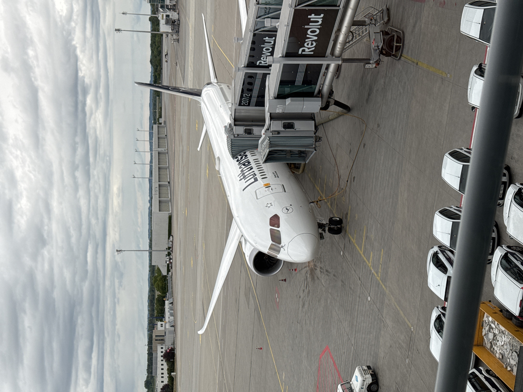 The image shows a Lufthansa Boeing 787-9 Dreamliner parked at Munich Airport (Flughafen München, MUC) in Freising, Germany. The aircraft is connected to two jet bridges advertising "Revolut". A row of white cars are parked adjacent to the aircraft. The sky is cloudy. The overall scene appears to be taken from an elevated position within the airport terminal.