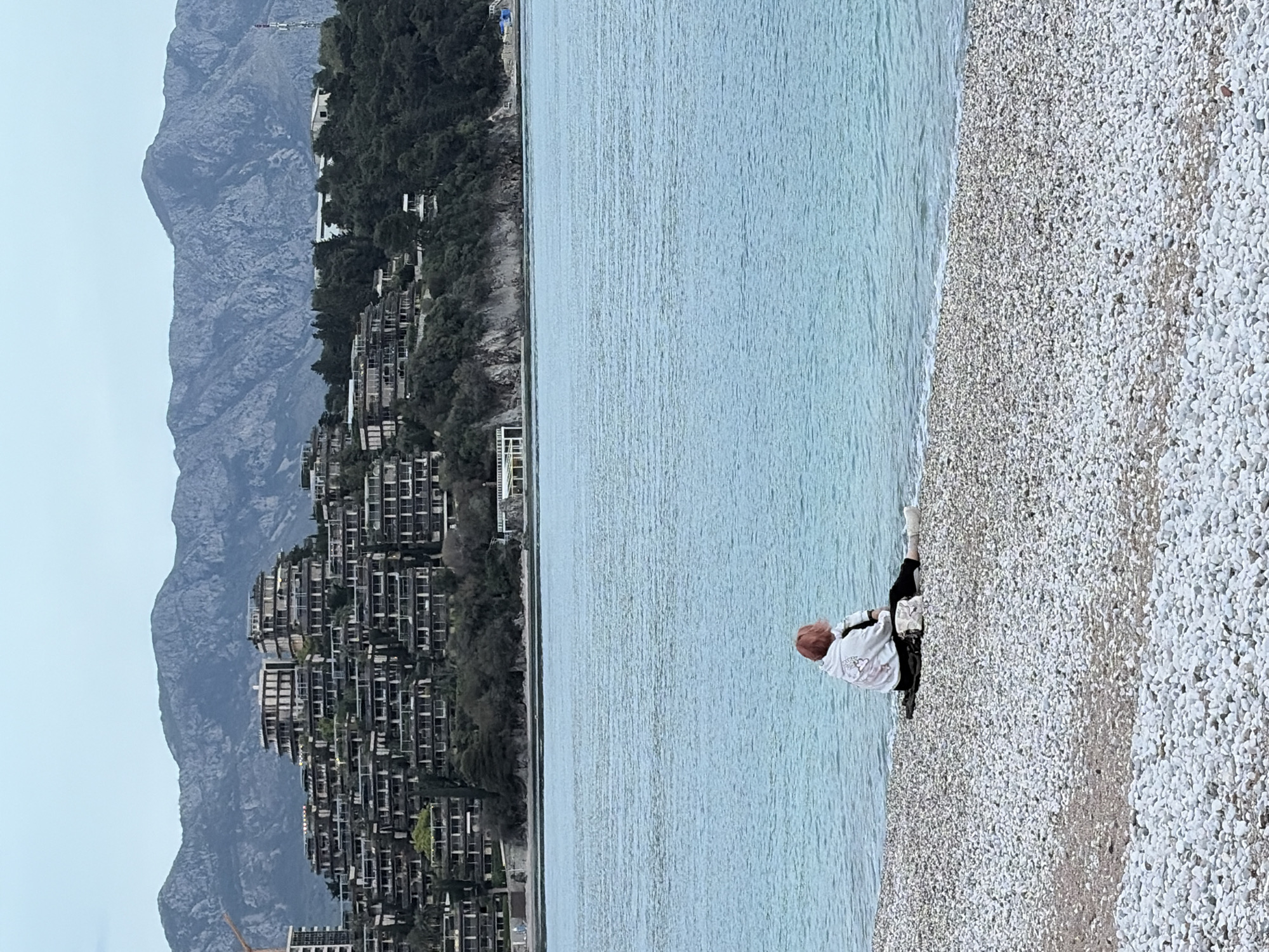 The scene depicts a beach in Budva, Montenegro. The environment includes a gravel beach transitioning into the blue-green sea, with a large mountain range and tiered buildings visible in the background. A single person, with reddish-brown hair, is sitting at the edge of the sea, possibly resting or relaxing. They are wearing black pants, white shoes, and a light-colored shirt or jacket with a graphic on the back. The buildings in the background appear to be residential or resort structures, built into the hillside. The sky is overcast, contributing to a muted color palette for the overall image.