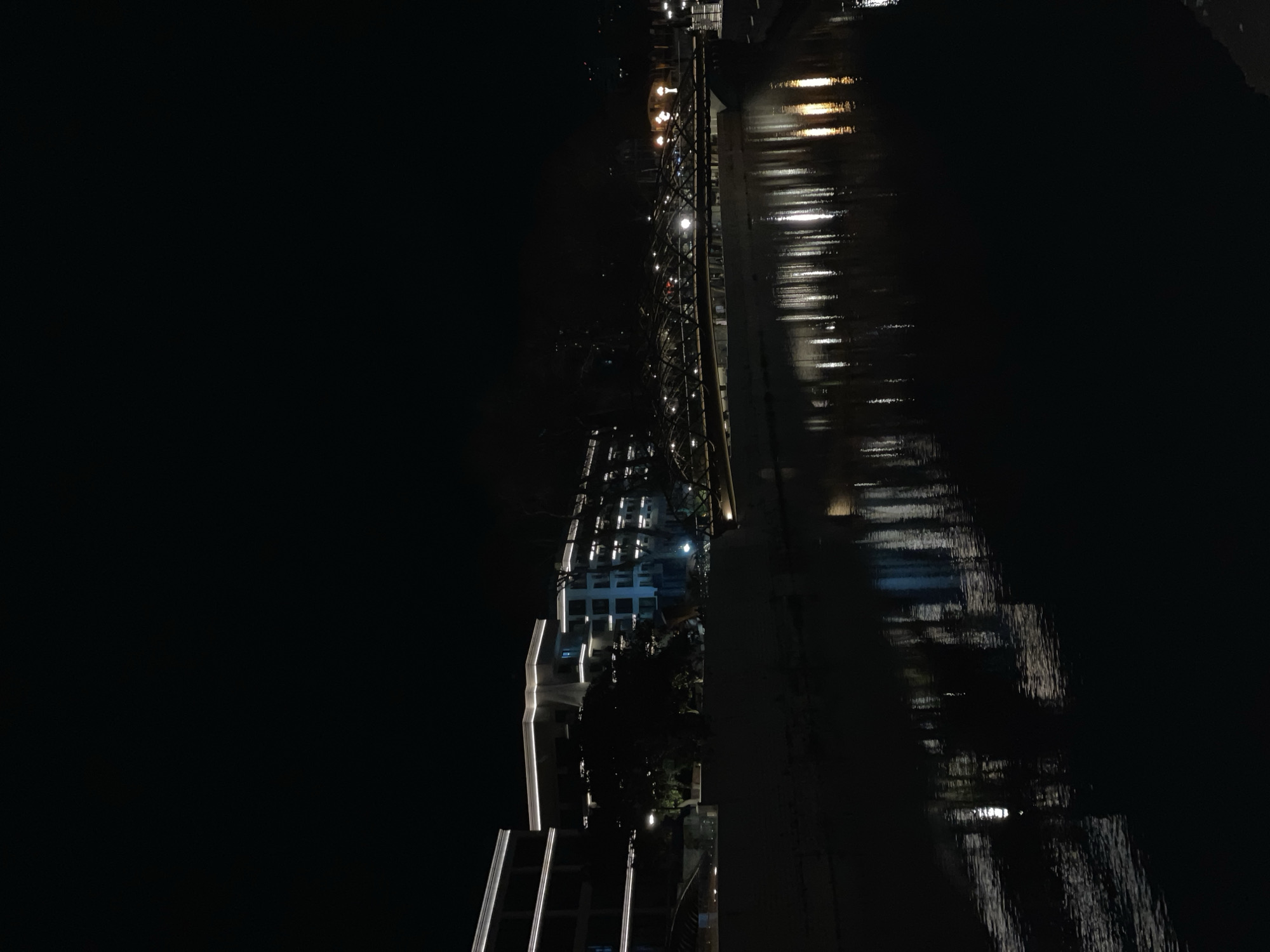The image displays a nighttime scene along the Mzymta River embankment in Adler, Sochi, Russia. The dark, calm river occupies the lower half of the frame, reflecting numerous artificial light sources as elongated vertical streaks. In the mid-ground, an illuminated pedestrian bridge with a dark, intricate arched lattice-like metal structure spans the river, featuring multiple uniformly spaced white lights along its length.
On the left riverbank, several multi-story modern buildings are visible, characterized by prominent white architectural lighting tracing their rooflines and edges. Dark trees and foliage are present along the embankments between the illuminated structures. Further down the river on the right bank, additional illuminated buildings and streetlights are discernible. The upper third of the image consists of a dark, starless night sky. No individuals or specific actions are visible within the frame.