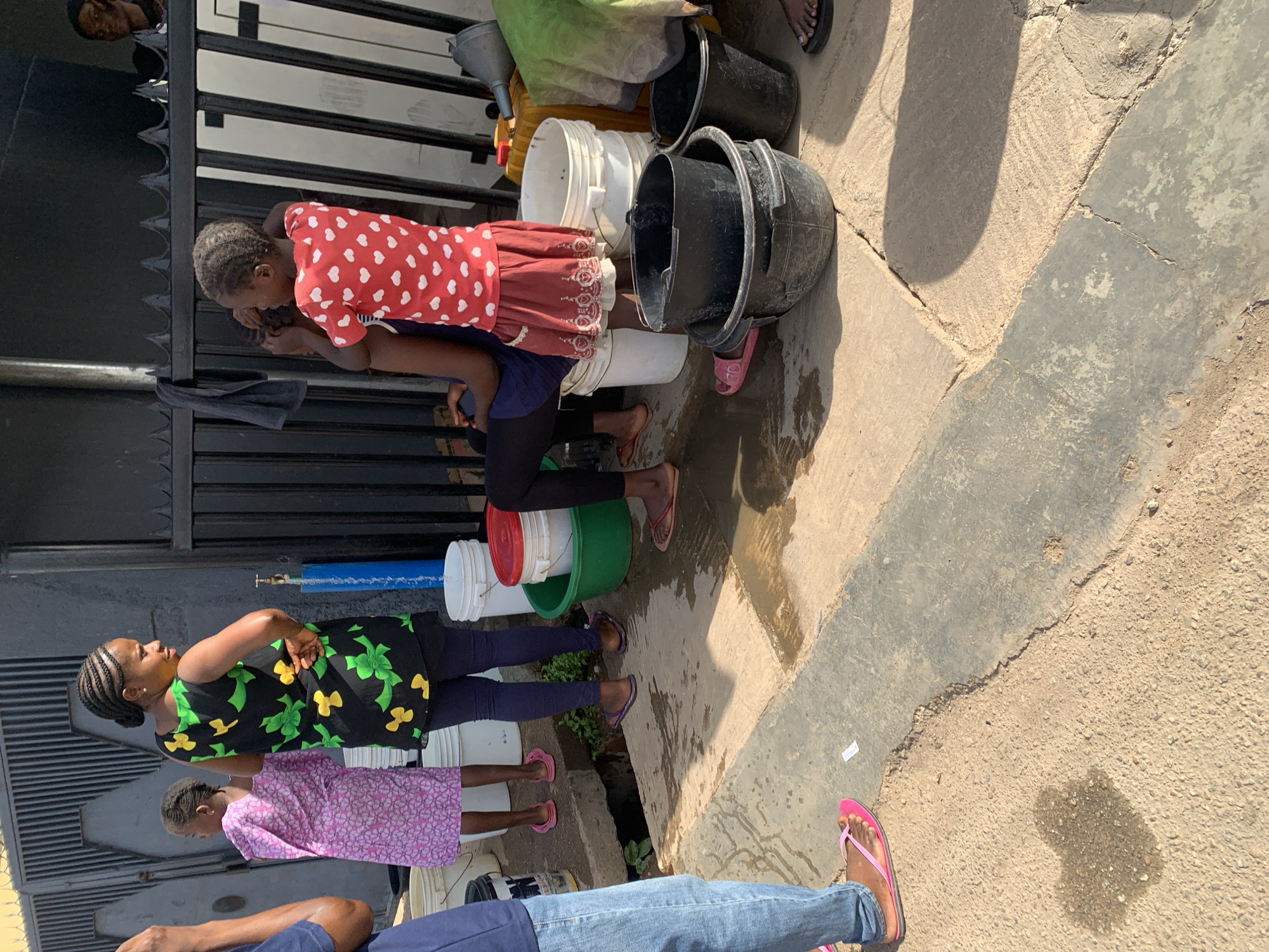 In Jos, Nigeria, a queue of individuals waits near a gate and wall to fill buckets with water. A girl in a red dress with white hearts is positioned over a filled bucket. A woman in a black dress with green and yellow flowers stands with her hands behind her back. Buckets of various colors, including white, black, and green, are visible, some containing water. The ground is concrete and has a water puddle on it.