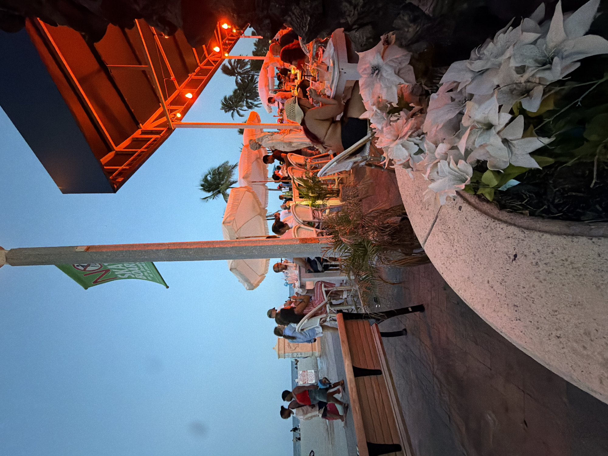 A twilight scene at an outdoor restaurant near the beach in Hollywood, Florida, USA. The sky is a clear, deep blue, transitioning into darker hues. A tall, light-colored pole with a green banner is visible on the left, featuring red lettering that reads "NO SKATEBOARD". In the foreground, a beige wooden bench sits on a textured grey paved area. To the right, the outdoor dining area is illuminated by warm red lights from an overhead structure, casting a glow on tables set with white umbrellas and chairs. People are seated at several tables, engaged in dining and conversation. Palm trees are sparsely visible against the sky. A stone planter with white artificial flowers is prominent in the lower right corner. In the background, beyond a low wall, is a sandy beach with a few people walking, and the ocean.
