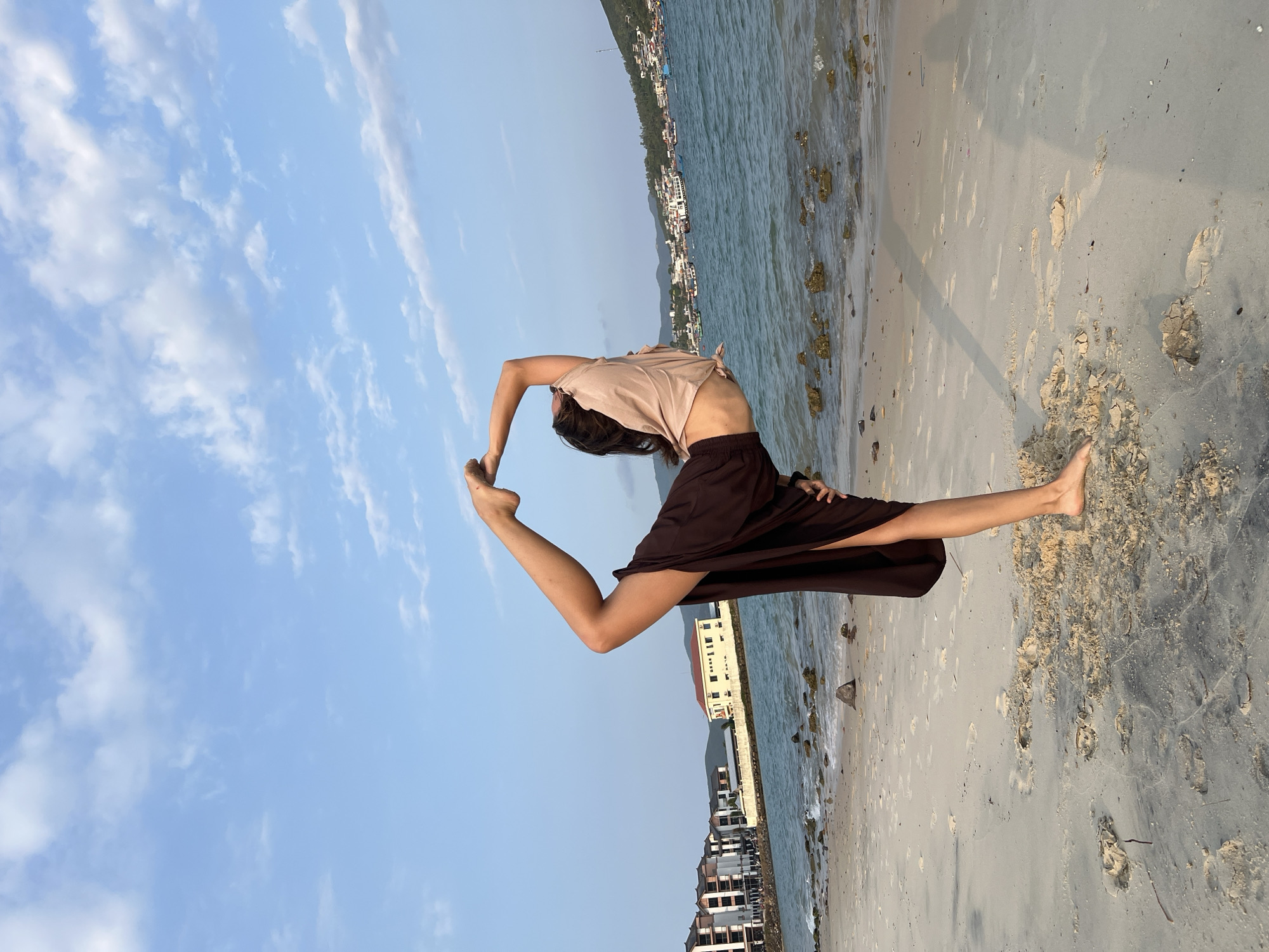 The image displays a person performing a challenging yoga or acrobatic pose on a sandy beach. The individual is balancing on one leg, with the other leg extended upwards and bent, while their arms reach back to grasp the raised foot, creating a distinctive "O" shape with their body. They are wearing a light-colored, possibly beige or tan, short-sleeved top and dark, flowing pants or a skirt.
The scene is a tranquil beach under a clear, light blue sky adorned with scattered white clouds. The foreground shows sandy ground with visible footprints and some patches of damp, darker sand. To the right, the ocean water, appearing a deep blue-green, washes gently onto the shore, where small rocks are visible at the water's edge. In the distance, along the coastline across the water, various buildings are nestled at the base of undulating hills or mountains.
The lighting suggests a soft, diffused sun, possibly late afternoon or early morning. The overall atmosphere conveys a sense of peace and natural beauty, combined with the focus and grace of the pose. No visible text is present in the image. The location is the Nam Natural Heritage Area Trang, Vietnam.