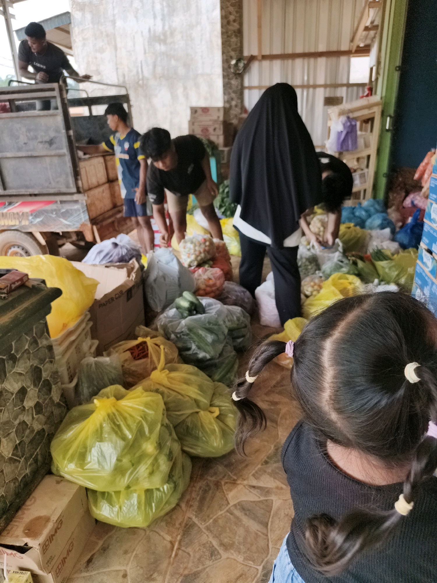 The image depicts a scene of people unloading produce from a truck, likely in a marketplace or distribution area. Three individuals are visible; one is standing on the truck bed, appearing to hand items down, while two others are on the ground, bent over and attending to the produce. A young girl with dark hair tied in pigtails is in the foreground, facing away from the camera, observing the activity.
The ground is tiled, and stacks of plastic bags filled with various vegetables and fruits are scattered around. Some bags are translucent yellow, while others are white, blue, and clear. A cardboard box labeled "PAPER CUP" and another partially visible box with "ULTRA MILK" text are also present. In the background, a woman in a black hijab is also handling produce. The setting appears to be an open-air structure with a corrugated metal roof and wooden supports, suggesting a warm climate. The overall atmosphere seems busy and functional, with a focus on the movement of goods. The time of day is unclear, but the lighting suggests it is daytime. There are no clear indicators of weather conditions.